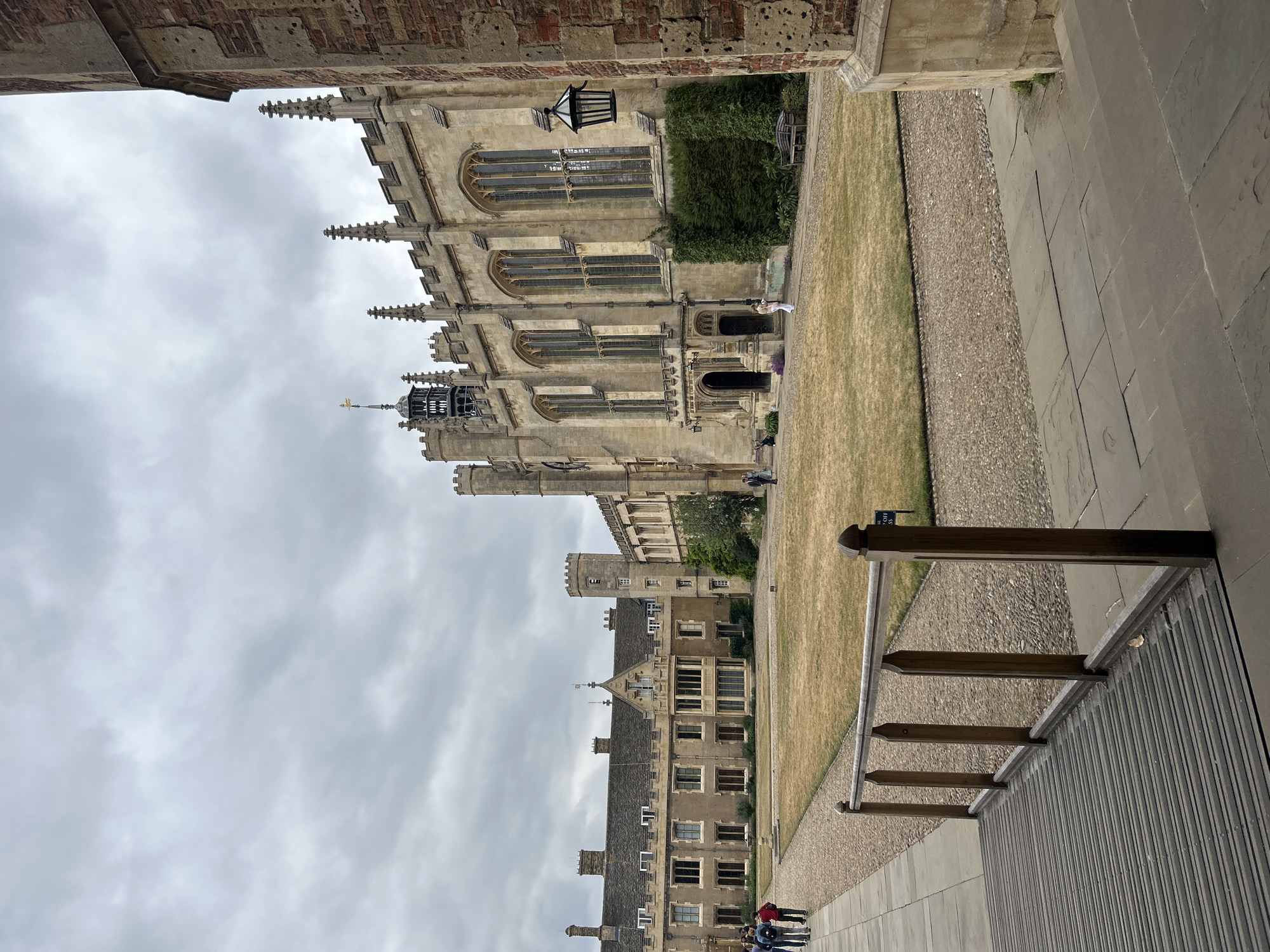 The image depicts a courtyard of a university, likely King's College in Cambridge, United Kingdom, given the architectural style. The sky is overcast with a high layer of clouds. The courtyard is paved with cobblestones in some areas and flagstones in others, with a central grassy area that appears dry and parched.
Dominating the right side of the image is a large, gothic-style building constructed from light-colored stone. It features numerous tall, arched windows, decorative crenellations along the roofline, and a prominent tower with turrets. A black lantern hangs on the exterior wall. To the left, another, older-looking building with a steeper roofline and numerous windows is visible. A smaller tower with a clock face is also present on this building.
In the foreground, a wooden railing with vertical posts and a horizontal top rail separates a raised wooden walkway or ramp from the rest of the courtyard. The walkway has a ribbed surface.
Several people are present in the courtyard. On the left, a group of four individuals, likely tourists or students, are walking away from the camera along the paved path. Further into the courtyard, near the entrance of the stone building, two individuals are standing, one appearing to be a woman in light-colored clothing. On the far right, near the base of the large stone building, a single person is walking. A wooden bench is situated against a wall covered in dense green ivy.
The overall impression is one of historical architecture and a calm, albeit somewhat muted, atmosphere due to the cloudy weather.