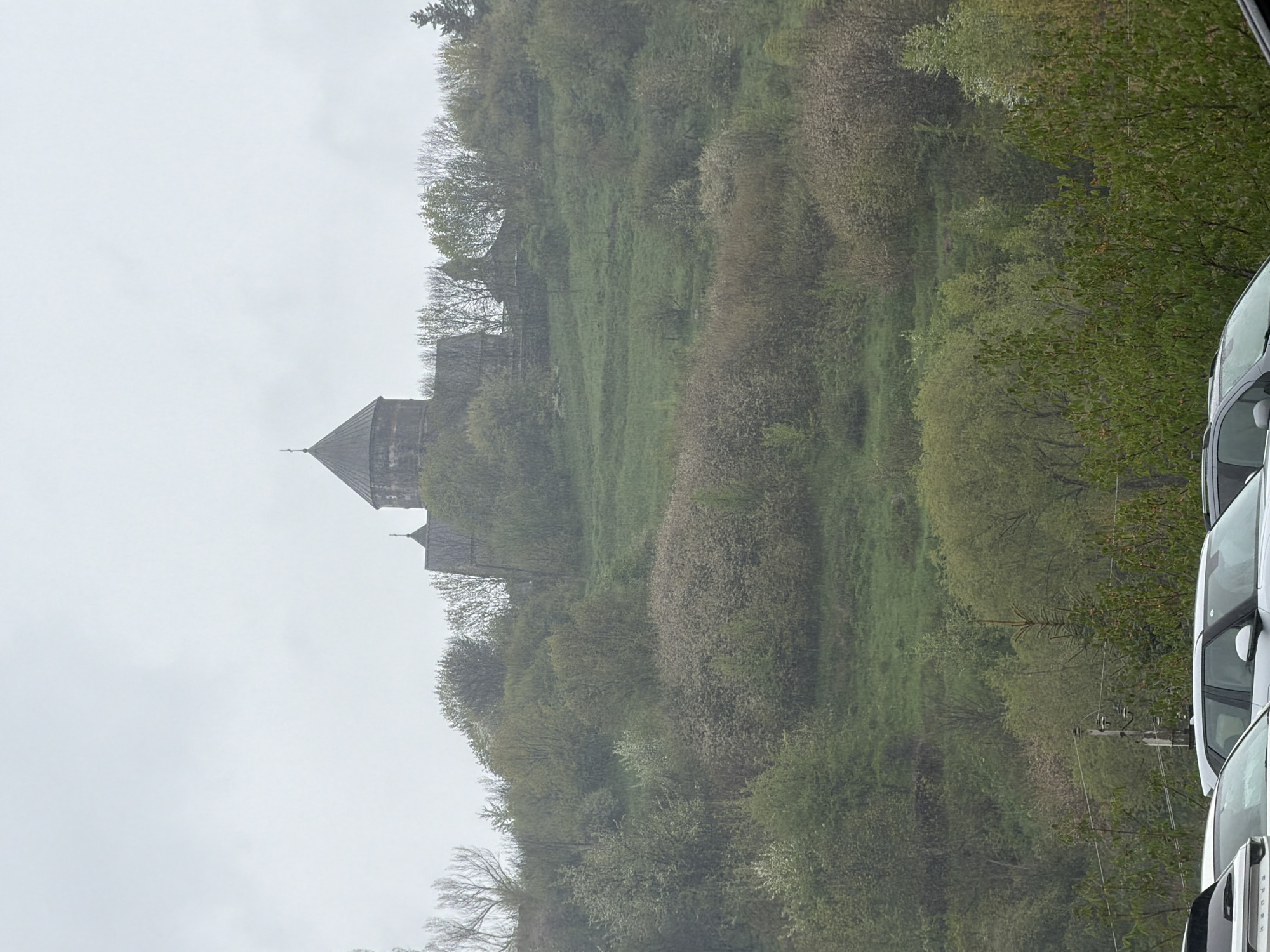 The image depicts a landscape with a prominent stone structure resembling a monastery or fortified building positioned on a hill. The building has a distinctive pointed roof. The hillside is covered with greenery, including trees and grass. The sky appears overcast. In the foreground, portions of several white vehicles are visible. The image is taken from what seems to be an observation point. The location is noted as Unknown, Unknown, suggesting the exact city or region is undetermined.