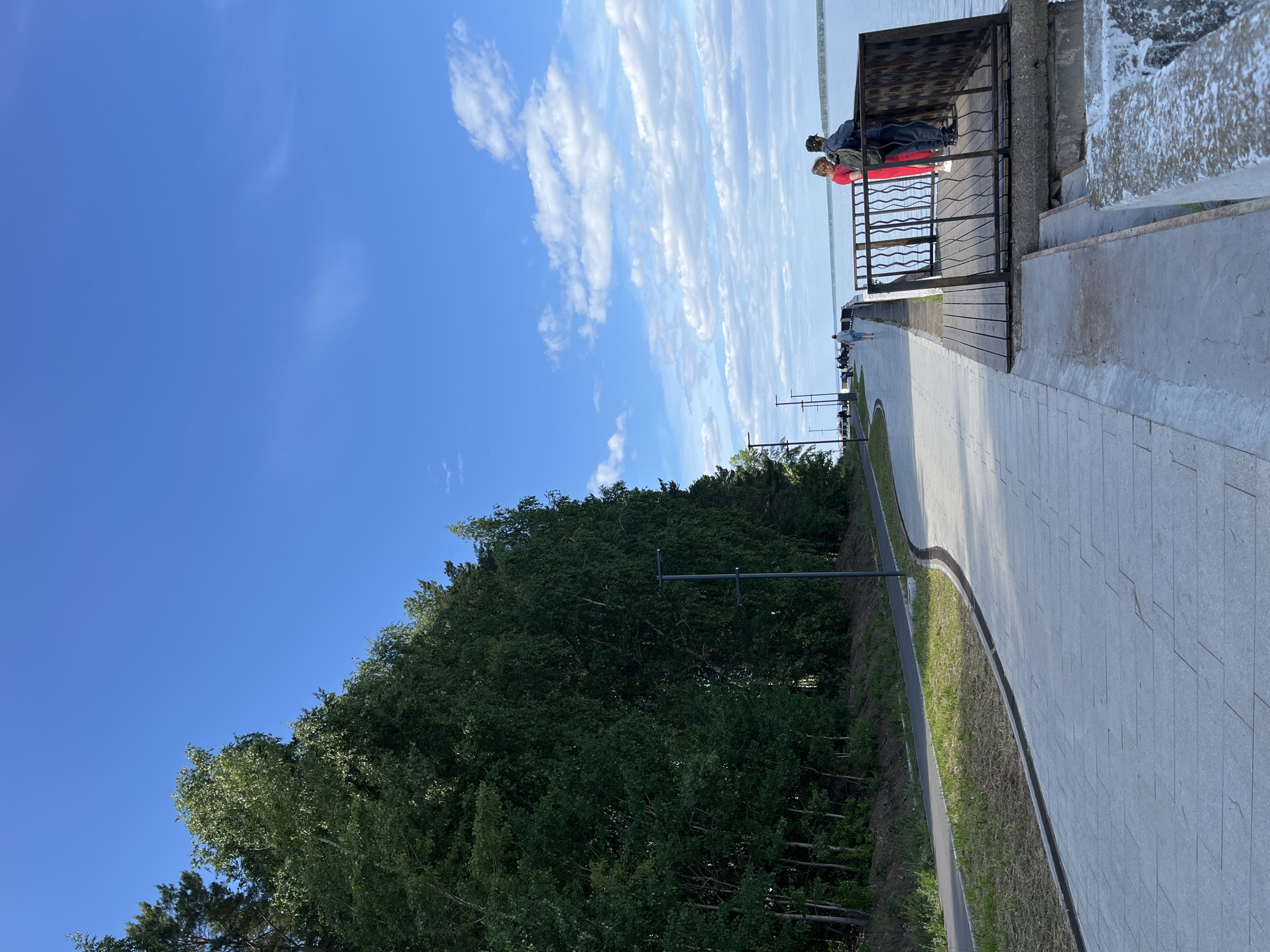 The image displays a daytime scene on the Chaikovsky Embankment, located in Chaikovsky, Perm Krai, Russia, likely bordering the Votkinsk Reservoir. The sky is clear blue with scattered white cumulus and stratocumulus clouds.
In the foreground and extending towards the midground on the left, a light grey paved promenade, constructed from rectangular pavers, spans the length of the scene. It is bordered on its left by a narrow dark drainage channel, a strip of green grass, and a separate, darker paved path, likely for cycling. Adjacent to these paths, a dense wall of tall, verdant trees, predominantly deciduous, forms a natural boundary, with their foliage extending high into the upper left quadrant. Several modern, dark grey streetlights are evenly spaced along the promenade's edge.
In the midground to the right, a dark wooden viewing platform with an intricate black metal railing extends over the water. Two individuals stand on this platform, both facing away from the camera and looking towards the water. One person wears a bright red top, dark trousers, and white shoes. The other person wears a dark jacket or top, dark trousers, and a dark cap, with a backpack visible. Several other indistinct figures are walking along the promenade further in the distance.
The background is dominated by a vast expanse of calm, light blue-grey water, stretching to the horizon where a faint, dark green landmass is visible.