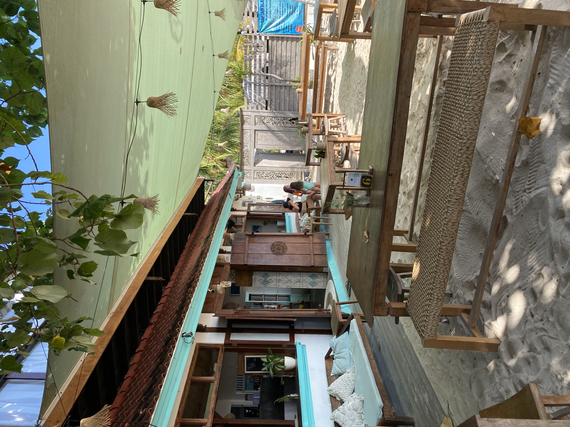 This image depicts the outdoor seating area of a beachfront cafe or restaurant, likely in Nusa Penida, Indonesia, given the tropical environment. The scene is set on a sandy beach, with several wooden tables and benches arranged under a large, pale green canopy that provides shade. Woven bamboo lamps hang from the canopy.
In the background, a building with a terracotta tile roof and turquoise trim is visible, featuring open doorways and windows. Some decorative wooden screens are placed near the building. A blue sign with text, possibly advertising services or rules, is mounted on a wooden fence.
Three women are seated at a table near the building, appearing to be engaged in conversation. One woman is wearing a black bikini top and hat, another a black bikini top and shorts, and the third is wearing a light blue swimsuit.
In the foreground, more wooden tables and benches are present. One table has a small sign with the number "6" and some small vases with greenery. A light blue cushioned bench with pillows sits on a wooden platform to the left. The sand is lightly rippled, suggesting a recent tide or wind activity. Lush green leaves from overhanging trees cast dappled shadows on the ground. The overall atmosphere suggests a relaxed, casual dining or lounging experience by the sea.
