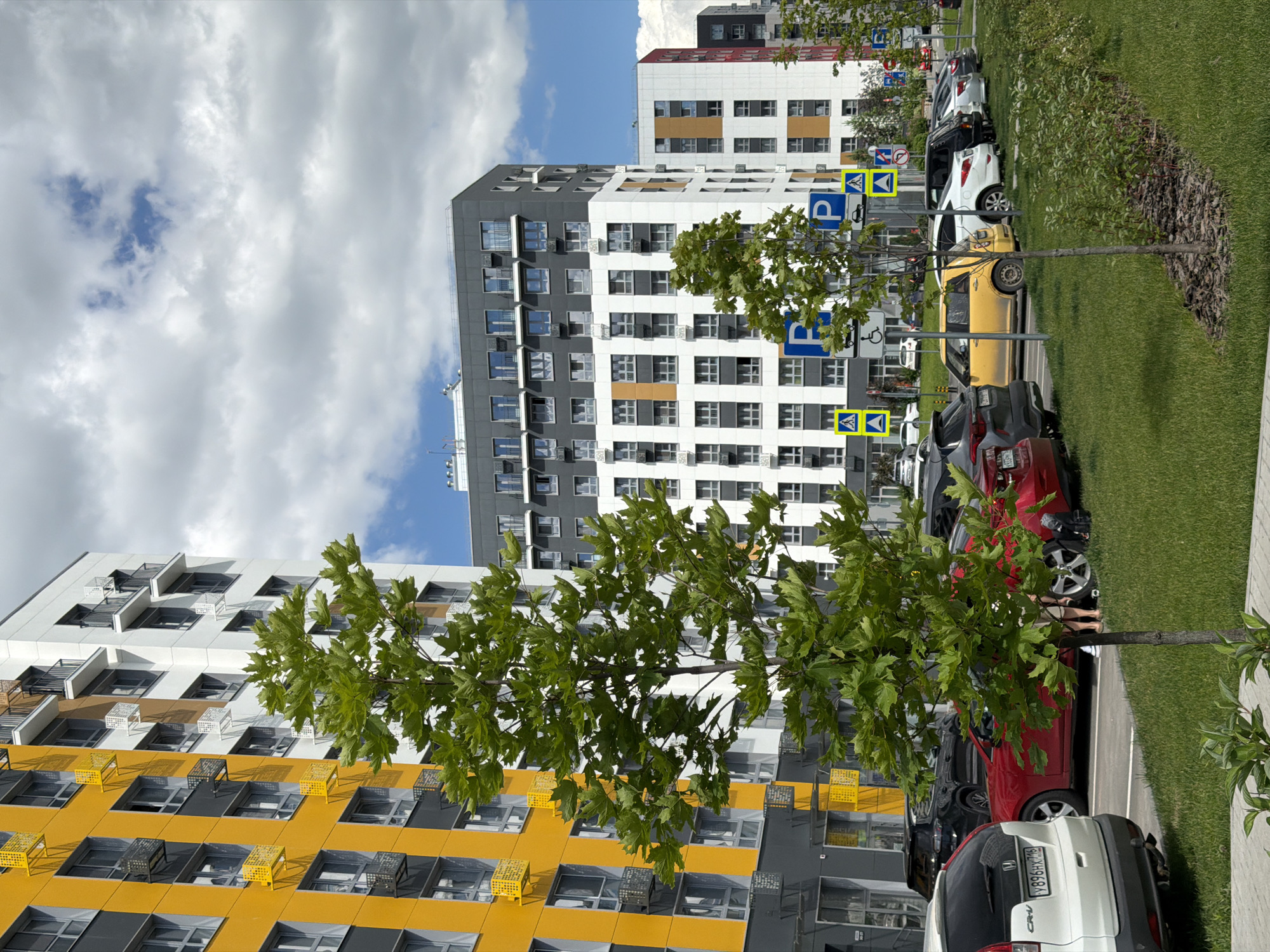 This is an eye-level, full shot taken on a cloudy day in the city of Kazan, Russia, depicting a modern residential complex. The buildings are a mix of yellow, white, and grey, with visible air conditioning units on many balconies. In the foreground, a young tree with green leaves stands on a grassy lawn, and a paved walkway is visible at the bottom. A line of parked cars, including a white SUV, a red sedan, and a yellow hatchback, are parked on the right side of the frame, along with several parking signs. The buildings are tall and multi-story, indicating a dense urban environment.