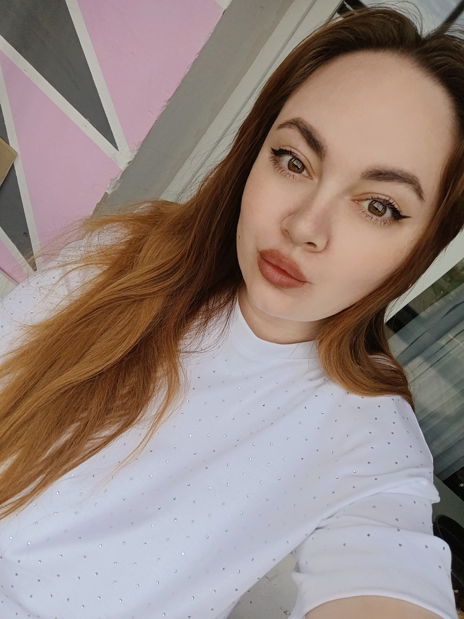 A selfie of a woman with long, reddish-brown hair. She is wearing a white T-shirt adorned with small, reflective rhinestones. She has brown eyes, styled eyebrows, and is wearing eyeliner. Her lips are done with a matte lipstick.
Behind her is a wall with painted geometric patterns of pink, grey, and white. The location is inferred to be Chaikovsky, Russia.