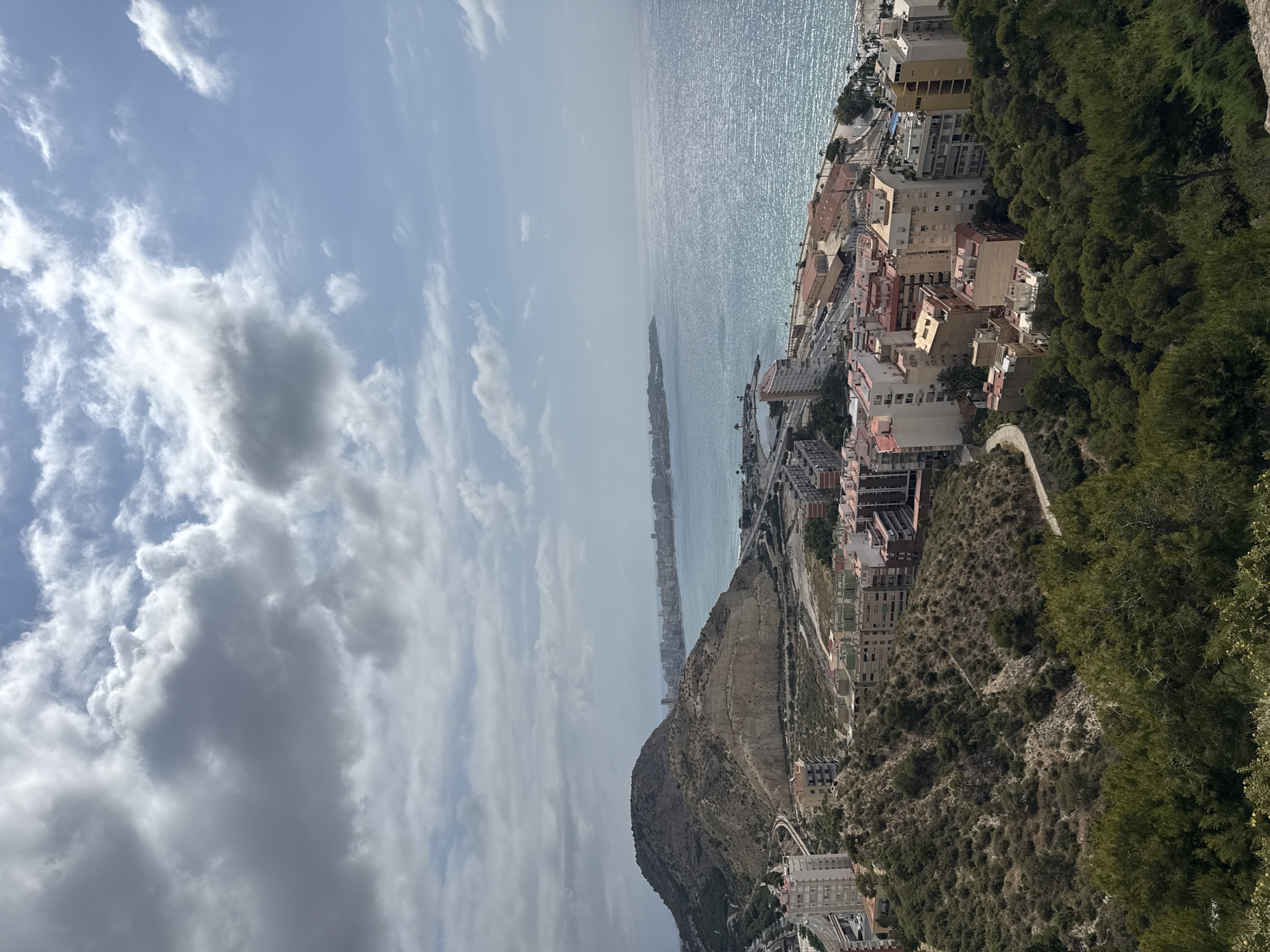 This is an aerial, wide-angle, outdoor photograph taken on a cloudy day. The scene captures a coastal city with a prominent hill in the foreground, overlooking the ocean. The hill is covered in dense, green foliage and scrub, with a winding path visible on its side.
Below the hill, the city unfolds with a cluster of multi-story residential buildings in various shades of white, beige, and red. These buildings are densely packed, with some appearing to be under construction or renovation, indicated by exposed concrete and scaffolding. A main road with some traffic runs along the coastline, separating the city from the sea.
The ocean is a vibrant turquoise, with sunlight glinting off its surface. The coastline stretches into the distance, lined with more buildings and a harbor area with small boats. In the far distance, a city skyline is visible against the hazy horizon. The sky is filled with dramatic, grey and white clouds, with patches of blue sky peeking through. The overall impression is one of a sunny day in a Mediterranean coastal city.