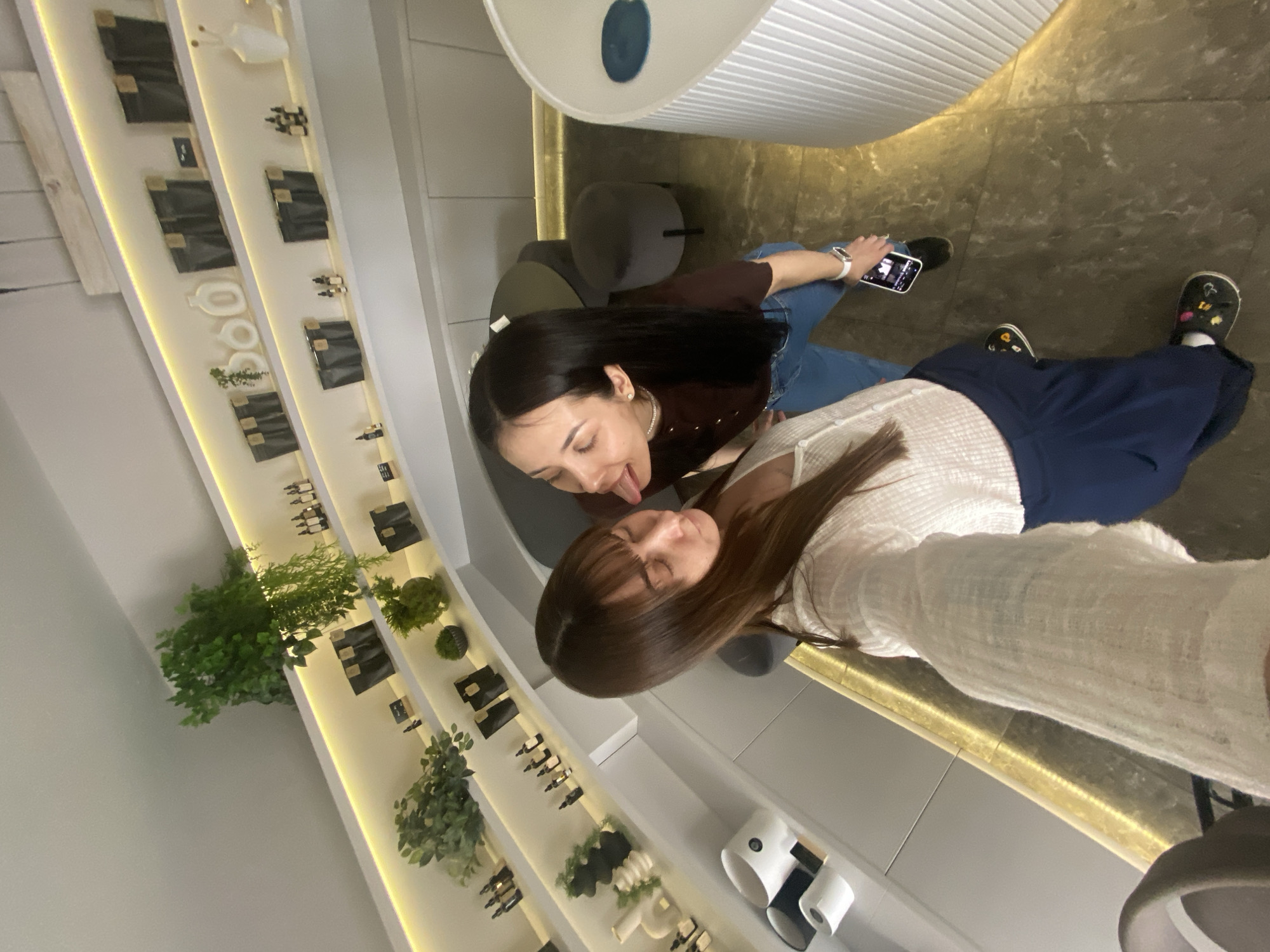 Two young women are in an indoor commercial space, likely a store or boutique. One woman is holding a smartphone and taking a selfie. The woman closest to the camera is wearing a light-colored, long-sleeved shirt and dark pants. The other woman is wearing a brown shirt and blue jeans. She is sticking her tongue out towards the first woman. They appear to be sitting on chairs, with additional seating nearby. Shelves with plants and retail items line the walls, illuminated with accent lighting. The location is Votkinsk, Russia.