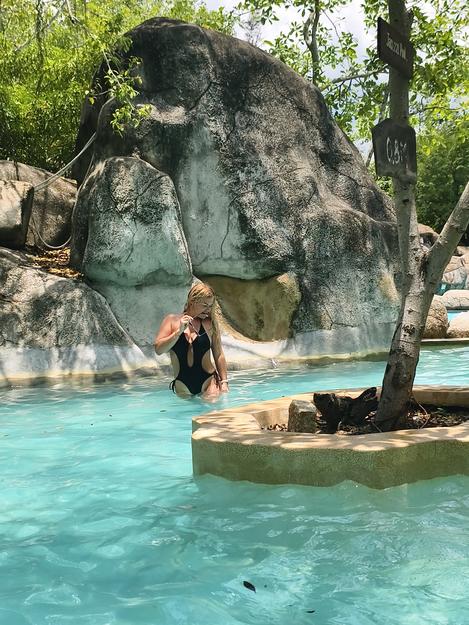 A woman stands in a pool of turquoise water at I-Resort Nha Trang in the city of Tay Nha Trang Ward, Vietnam. She has blond hair and is wearing a black one-piece swimsuit. She is holding sunglasses near her mouth. Behind her is a large, gray rock formation. A sign that reads "Jacuzzi Pool" is visible on a tree to the right. The water is clear, and the pool is surrounded by rocks and trees.