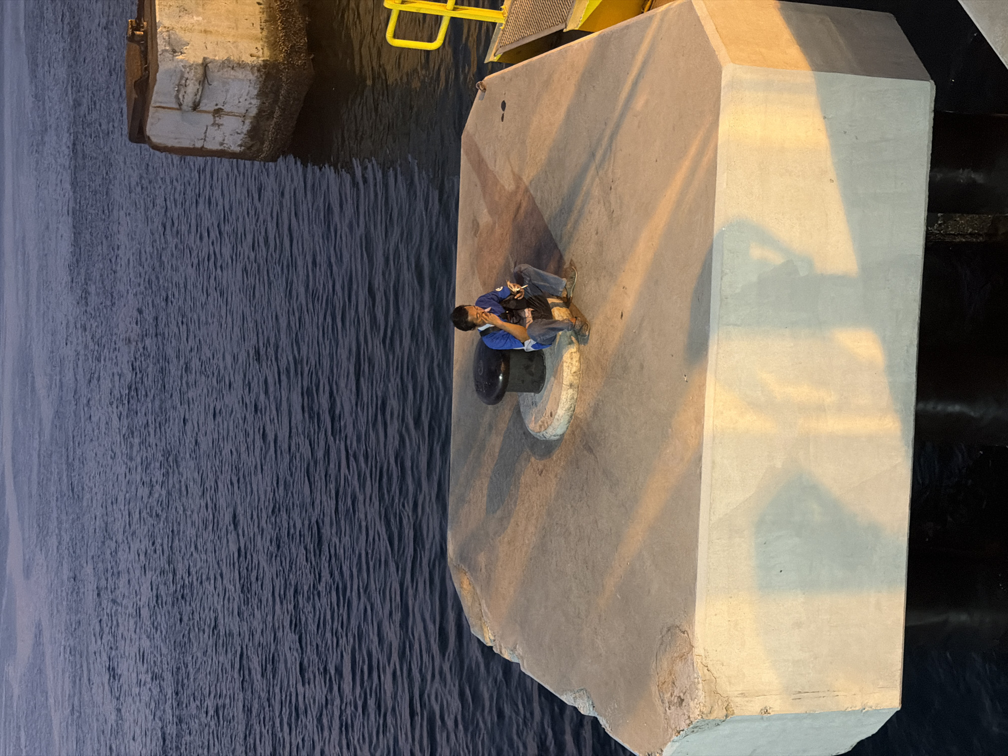 A man sits on a concrete platform by the water in Melaya, Indonesia. He is wearing a blue shirt and jeans, and he is holding a cigarette and a phone. The concrete platform is illuminated by artificial lights, casting long shadows on the ground. The water is dark and calm, with a gentle ripple. In the background, a concrete structure juts out of the water, and a yellow ladder and railing are visible on the right side of the image. The scene appears to be a quiet, contemplative moment by the sea.