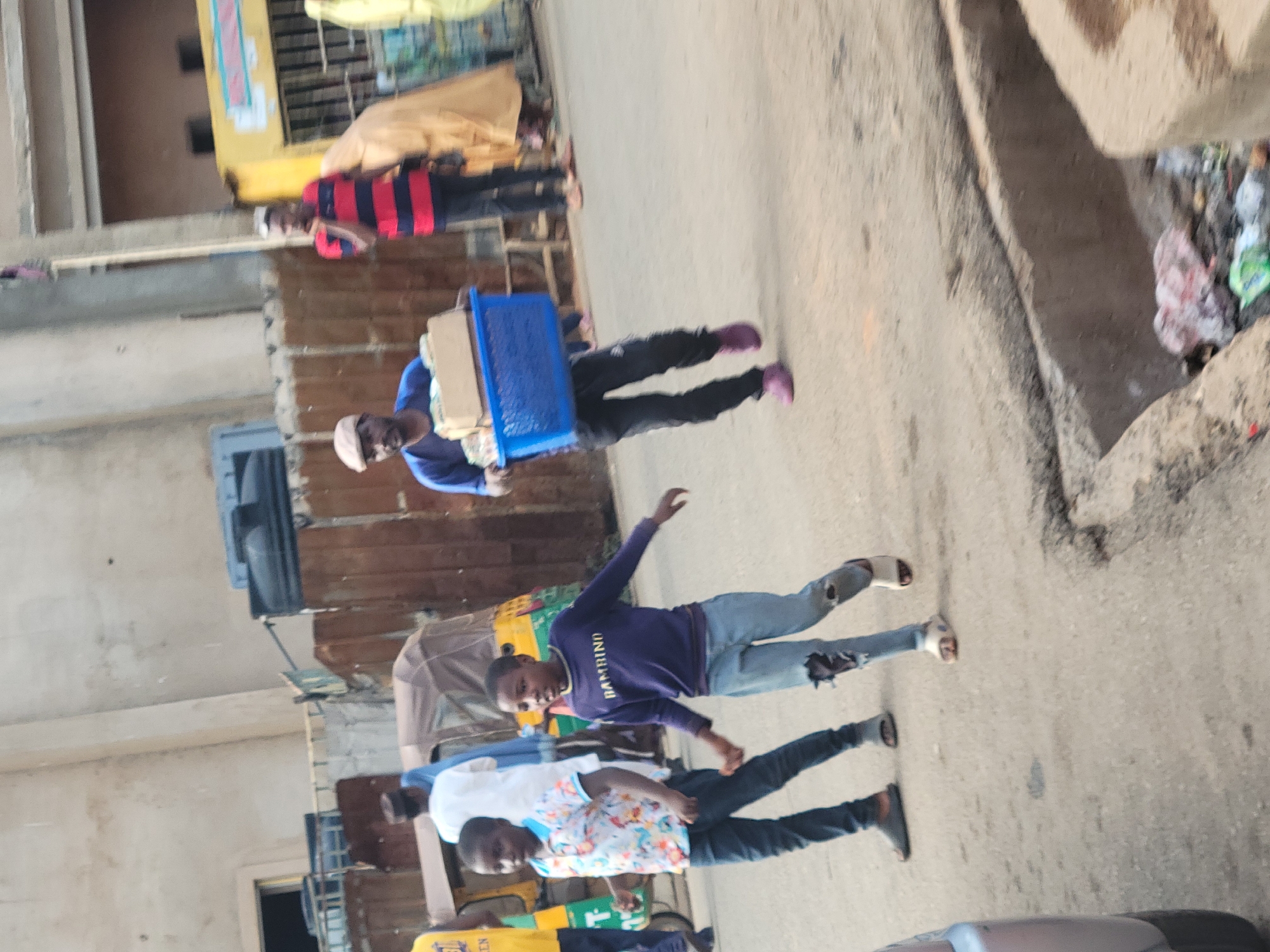 The image depicts a street scene in Jos, Nigeria. Several people are walking along a paved road. One man carries a blue basket with items inside. Two boys, one wearing a "Bambino" shirt, are mid-stride. Another man wears a floral shirt. Buildings line the street, including one with a yellow sign. There is trash on the side of the road.