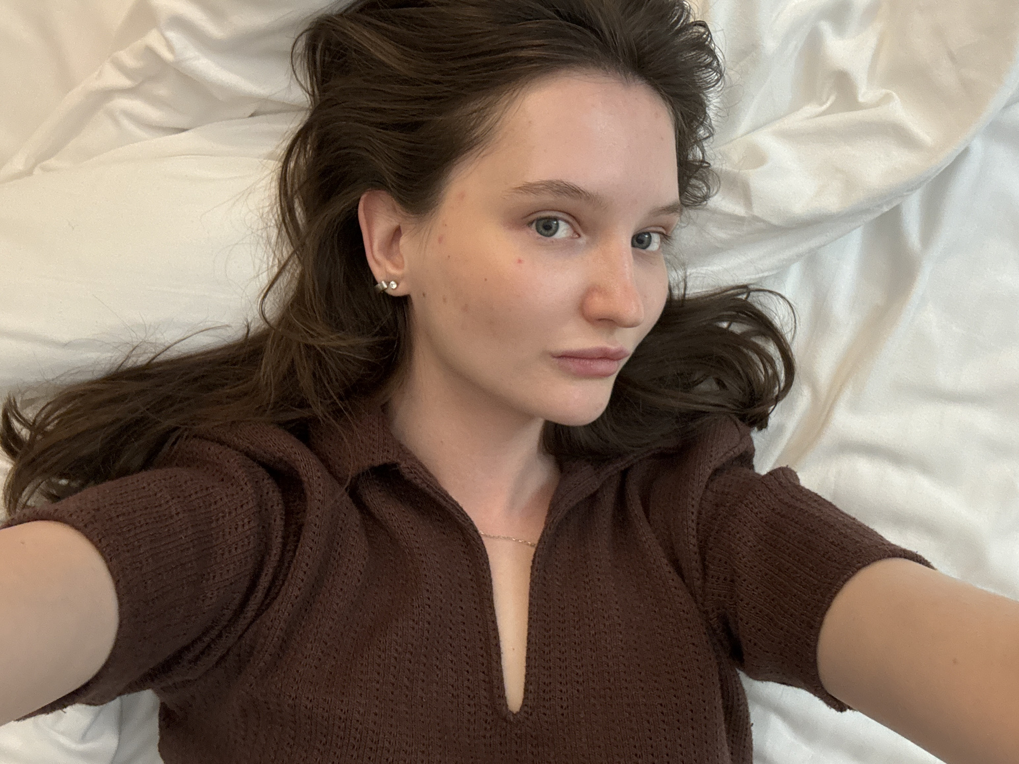 A young woman with long, dark brown hair lies on a white, creased bed. She is wearing a brown, knitted top with a cutout at the chest. Her head is tilted to the left, and she is looking directly at the camera with light blue eyes. Her skin is pale and clear, with a few scattered blemishes. She has a delicate silver chain necklace visible against her skin. The image appears to be taken indoors, possibly in a bedroom. There is no information available regarding the specific location in the city of Журавлево (кемеровский), Russia.