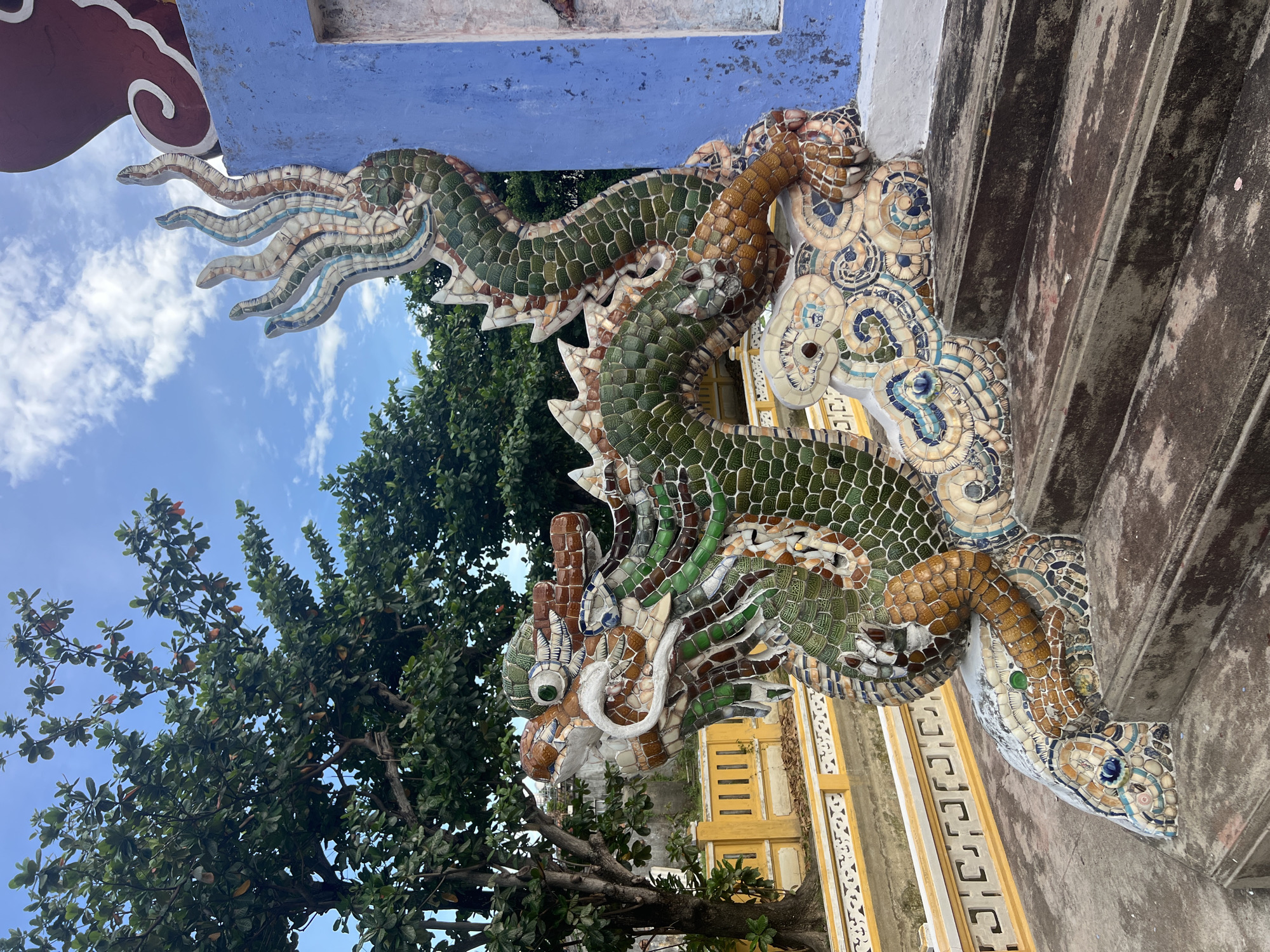 The image shows a decorative ceramic dragon sculpture at the entrance of a building in Hoi An, Vietnam. The dragon is vividly colored with green, brown, and blue mosaic tiles, and it appears to be coiled on a staircase. Behind the dragon, a vibrant blue wall can be seen, along with a glimpse of a red and white decorative element at the top. Lush green trees frame the scene under a bright blue sky with scattered white clouds. A yellow tiled fence with a decorative pattern is visible in the background. The overall impression is one of cultural and artistic richness.
