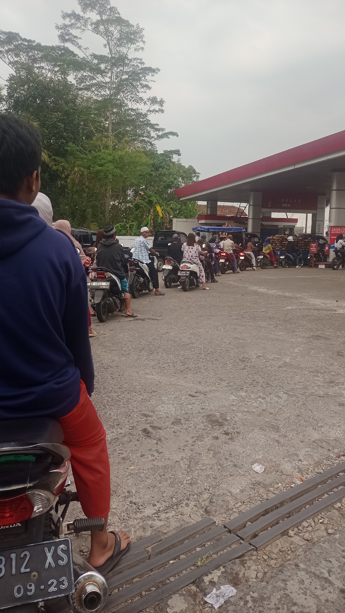 The image shows a queue of motorcycles and their riders at a gas station in Karyamandala, Indonesia. The gas station has a red and white canopy. The ground is paved with asphalt and there are some trees and foliage in the background. The sky is overcast. People are dressed in casual clothing, some are wearing religious headwear. The overall scene depicts a typical day at a local gas station in Indonesia.