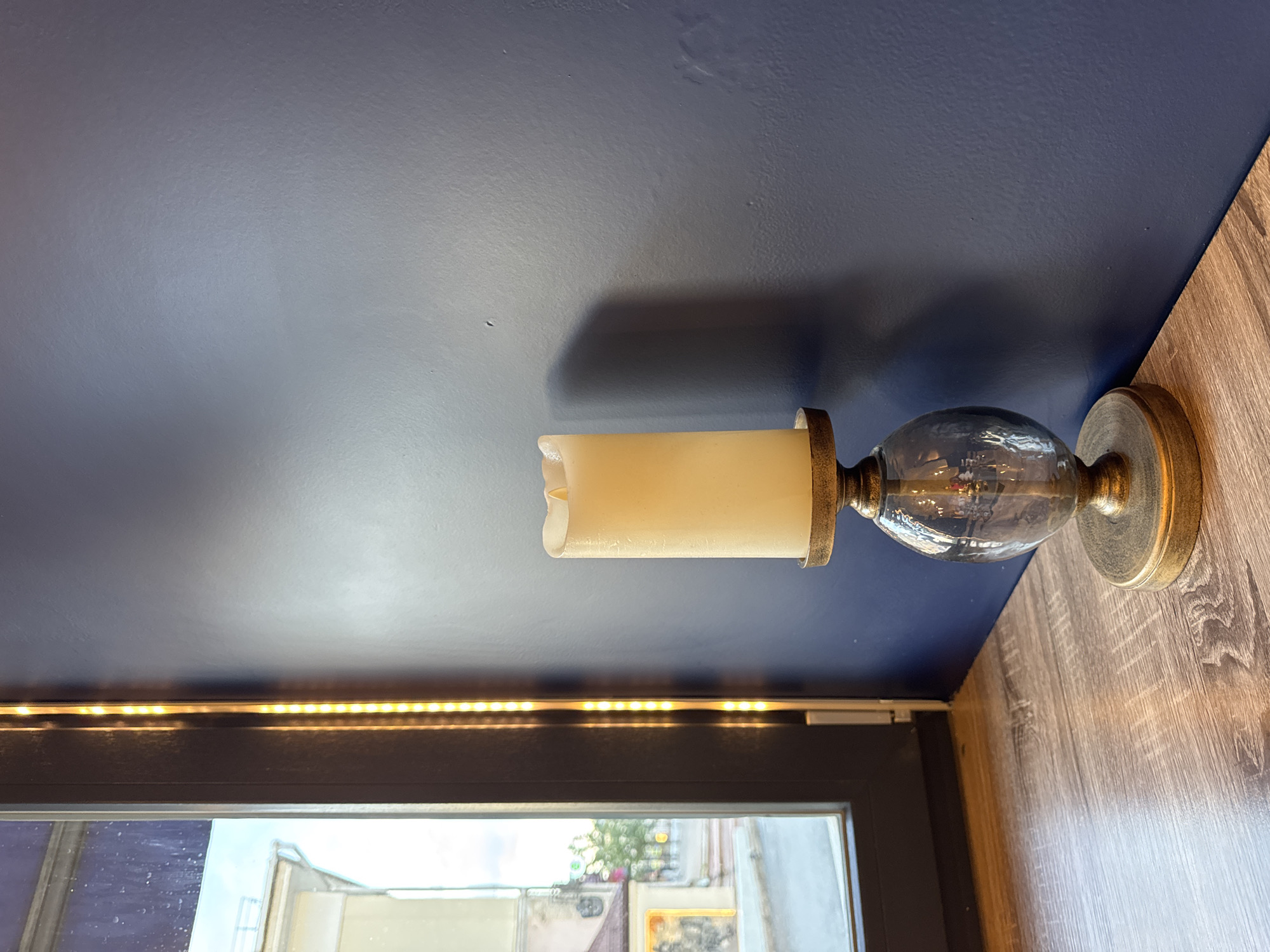 The image depicts a decorative candle on a wooden surface, positioned against a dark blue wall. To the left of the candle is a window with a view of a building and a street, suggesting an urban setting. The scene is illuminated by natural light coming from the window and an LED strip light running vertically along the window frame. No people are visible in the image. The overall impression is of an indoor scene with a view of the exterior. The location is a city, specifically Kazan, Russia.