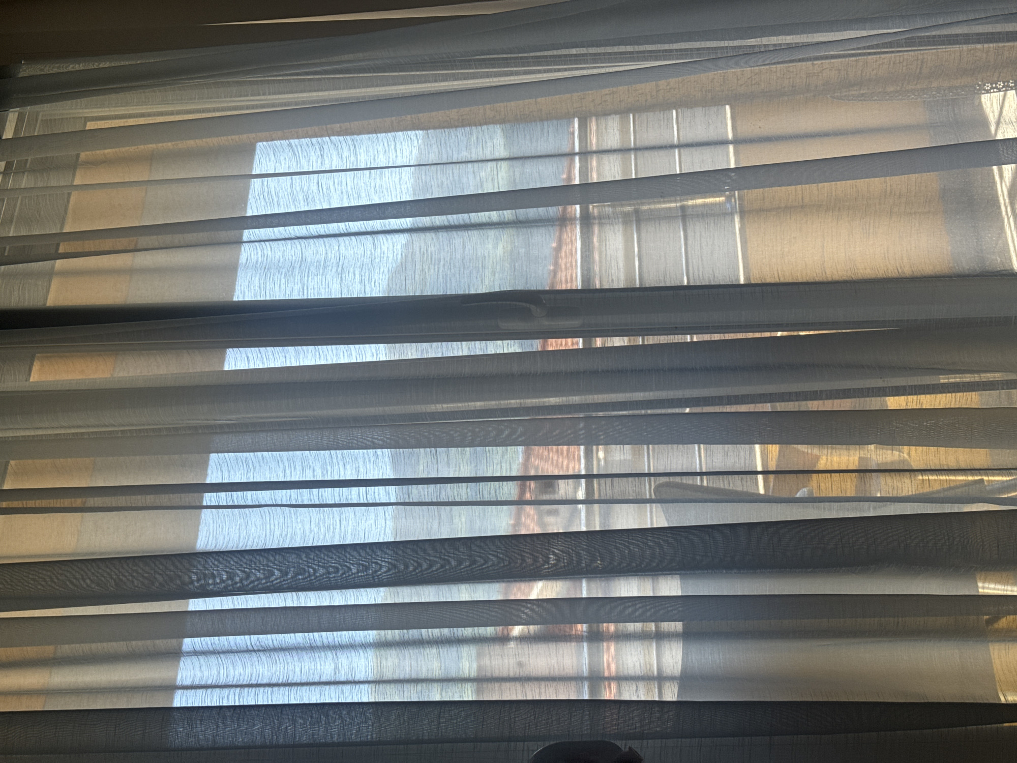 The image depicts a close-up view through a set of horizontal blinds. Through the slats, there is a blurred outdoor scene. The colors suggest daylight. Based on the crop images, a brick building is visible through the blinds. Based on the limited information, a precise location in Budva, Montenegro cannot be determined.