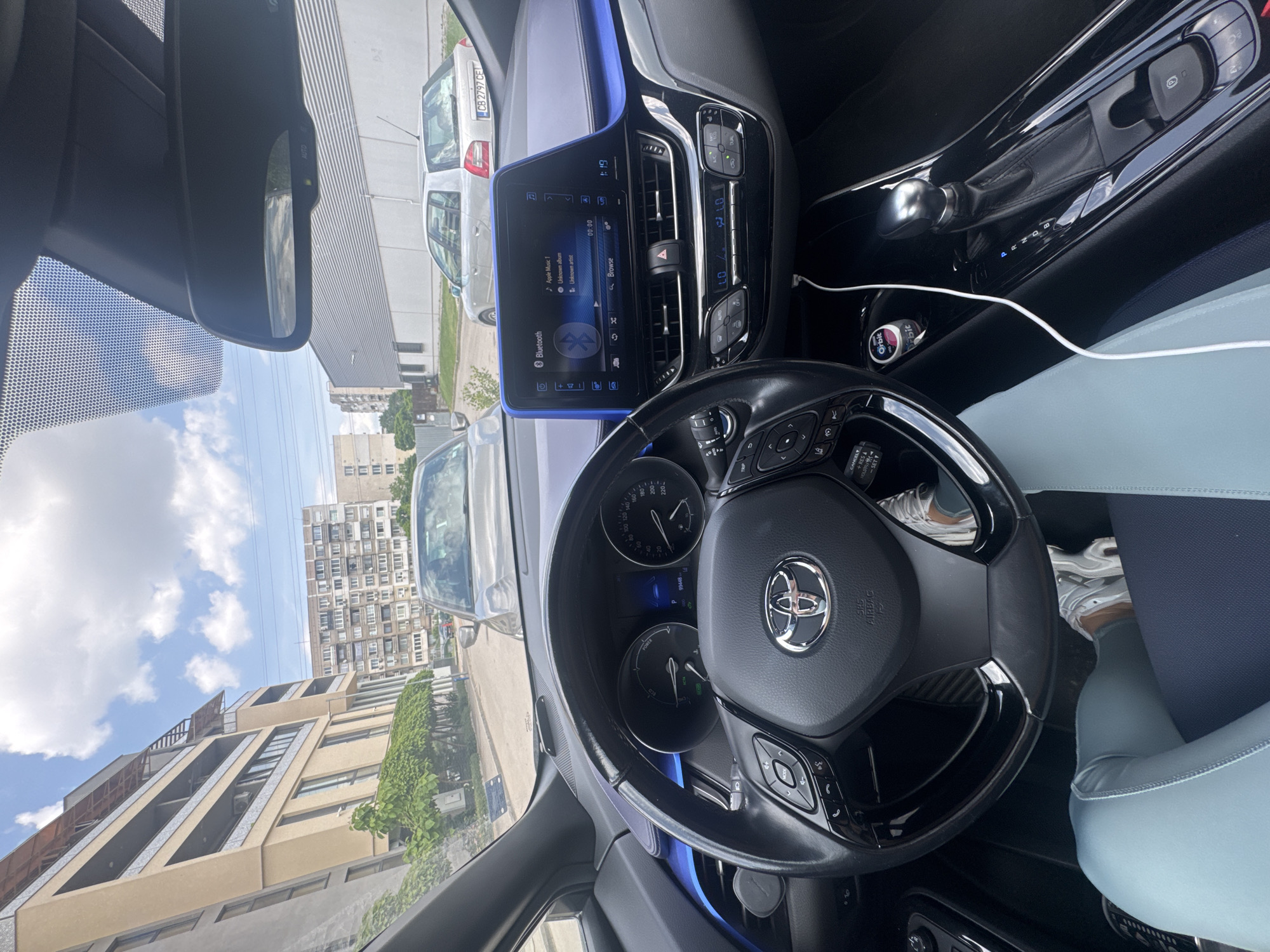 The image shows the interior of a Toyota C-HR, focusing on the dashboard, steering wheel, and a partial view through the windshield. The infotainment screen displays "Apple Music 1" with an unknown album and artist and Bluetooth connection. The driver is wearing athletic pants and sneakers. Outside the car, the view shows urban buildings, parked cars (one with the license plate "CB 2797 CE"), and a partially cloudy sky. Given the license plate and the architectural style of the buildings, the location is likely Sofia, Bulgaria.