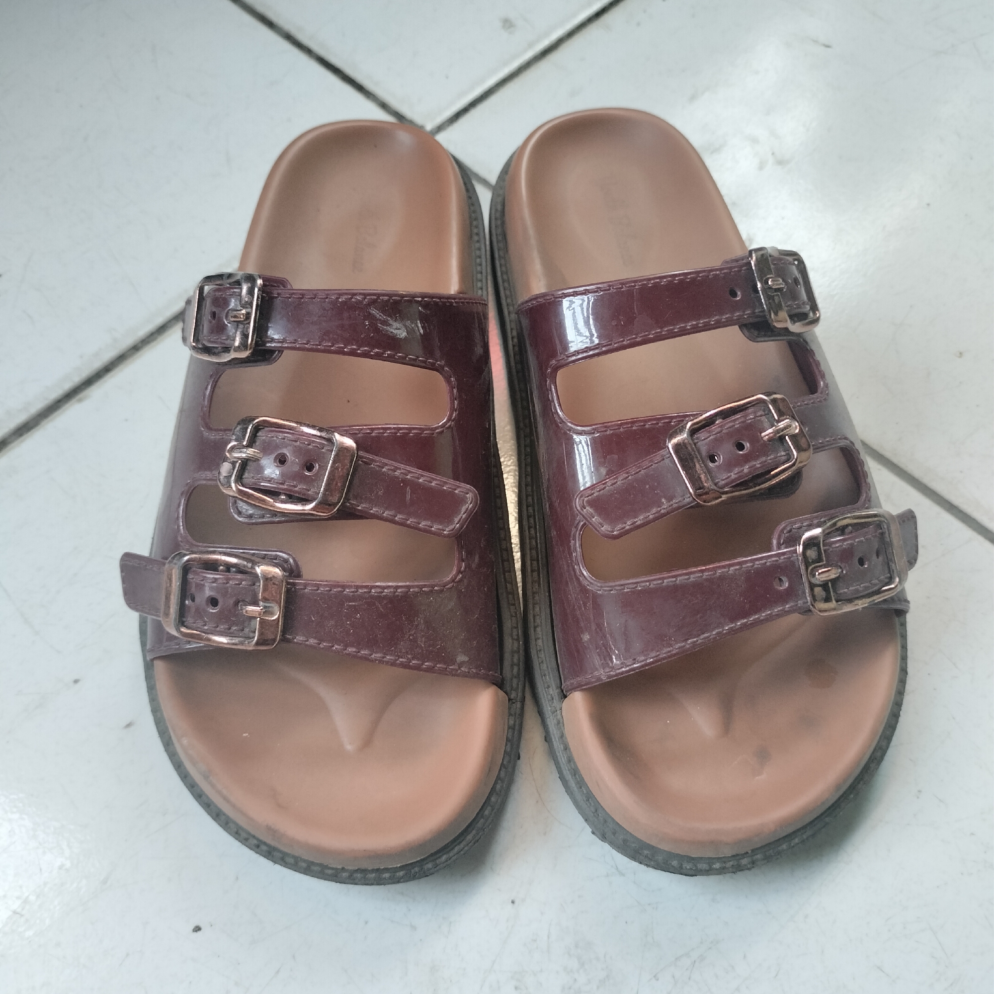 The image shows a pair of women's burgundy patent leather slides with three adjustable buckles on each strap, resting on a tiled floor. The sandals appear to be worn and have some scuff marks and dirt. The floor is made of square white tiles with visible grout lines. The lighting suggests it is daytime. The scene is indoors, likely a residential setting in Tangerang, Indonesia, given the context provided, although no specific landmarks or identifying features confirm this location.