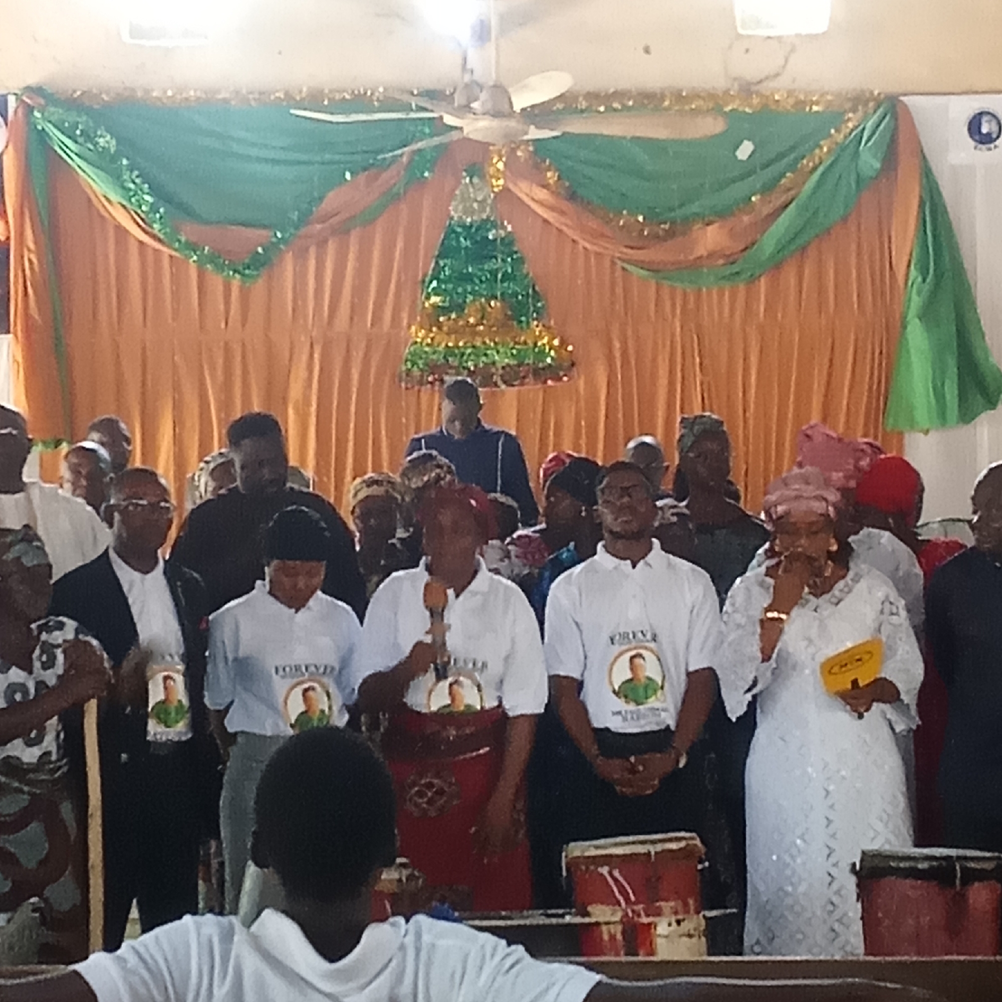 The image shows a gathering of people in an indoor setting, potentially a church or hall, adorned with green and orange drapery and a decorative element resembling a Christmas tree. The location is Unknown, Unknown. A group of people stand in the foreground and background. The crowd consists of males and females. The women are wearing gowns and headscarves of various colors. Some of the people in the foreground wear white T-shirts with the word "FOREVER" and a portrait on them. At least two people hold microphones. Drums are positioned in front of the crowd.