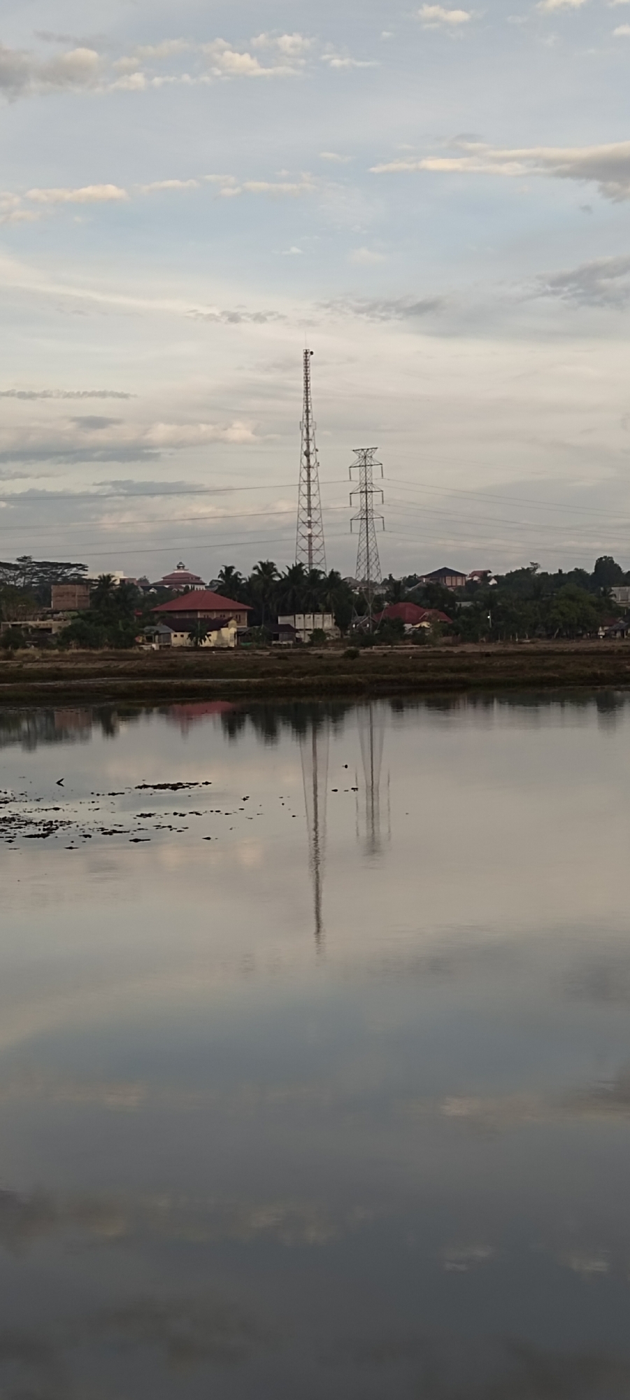 The image depicts a tranquil water body in the foreground, likely a rice field or a lake, with its surface reflecting the sky and the scene beyond. Scattered debris is visible on the water's surface. In the midground, a strip of land separates the water from a residential area. This area features houses with red-tiled roofs, interspersed with palm trees and other vegetation. Behind the houses, two prominent electrical towers stand tall, with one being a taller, slender telecommunications tower and the other a wider, more robust power transmission tower. Power lines extend from the latter. The background consists of a sky filled with scattered clouds, illuminated by soft, diffused light, suggesting either early morning or late afternoon. The overall atmosphere is serene and rural. The presence of telecommunications and power transmission infrastructure suggests it's an area with access to modern utilities, possibly a developing outskirts of a city. Given the specific infrastructure and the typical landscape found in Lhokseumawe, Indonesia, this scene is likely situated in the outskirts of the city, potentially near agricultural lands that are being developed for residential or industrial purposes.