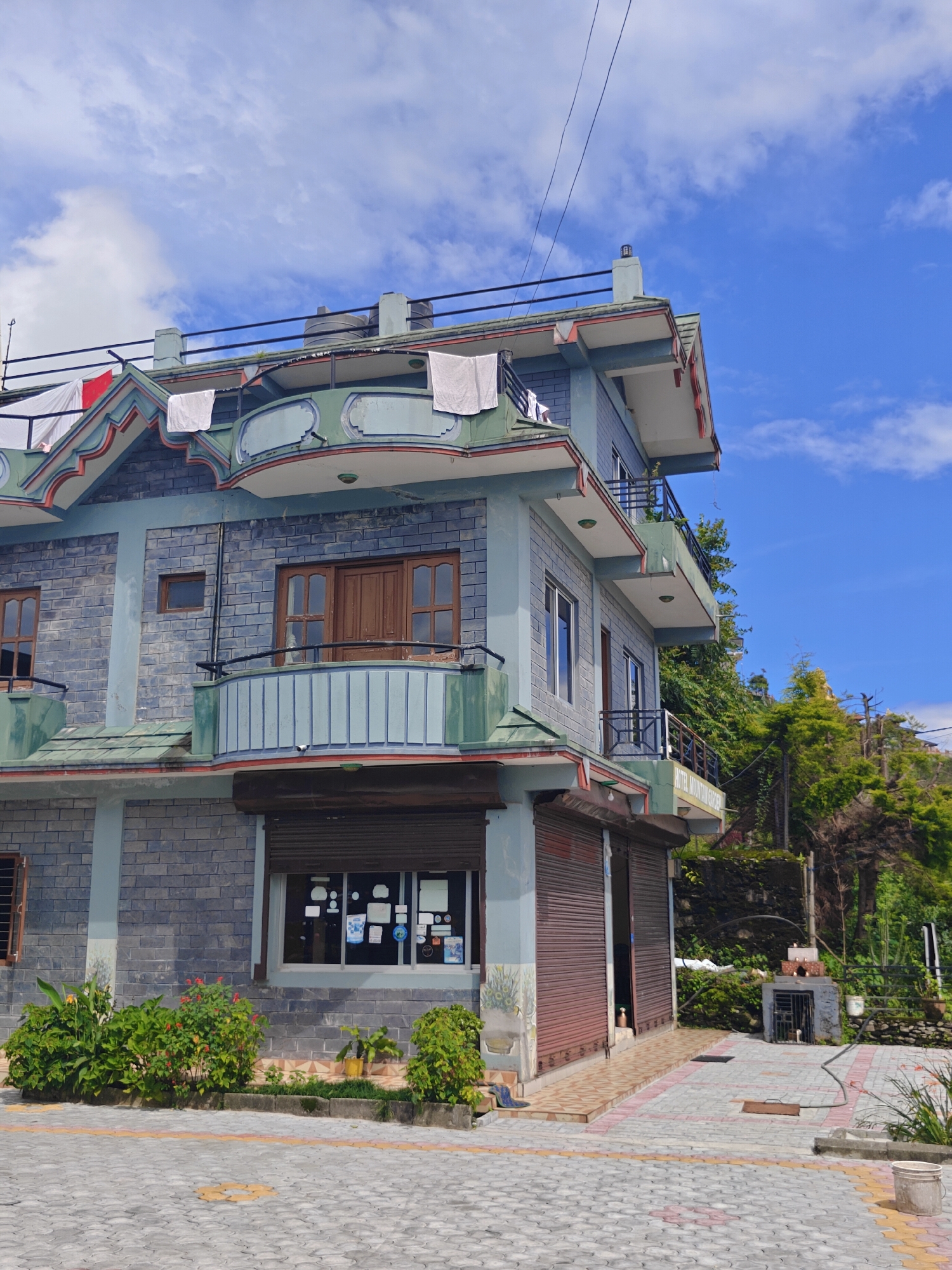 The image shows a three-story building with a blue-grey brick facade and green trim, located in Pokhara, Nepal. The building has multiple windows and balconies, and a rooftop with water tanks and laundry hanging out to dry. The ground floor appears to be a shop with a rolled-down metal shutter. In front of the building, there is a paved area with a floral garden and some potted plants. In the background, lush green trees and a hillside are visible under a bright blue sky with scattered clouds. A small, brick structure with a metal grate, possibly an outdoor oven or grill, is situated to the right of the building, along with some gardening tools and a bucket. The scene suggests a commercial or residential area in a hilly, possibly tropical, region.