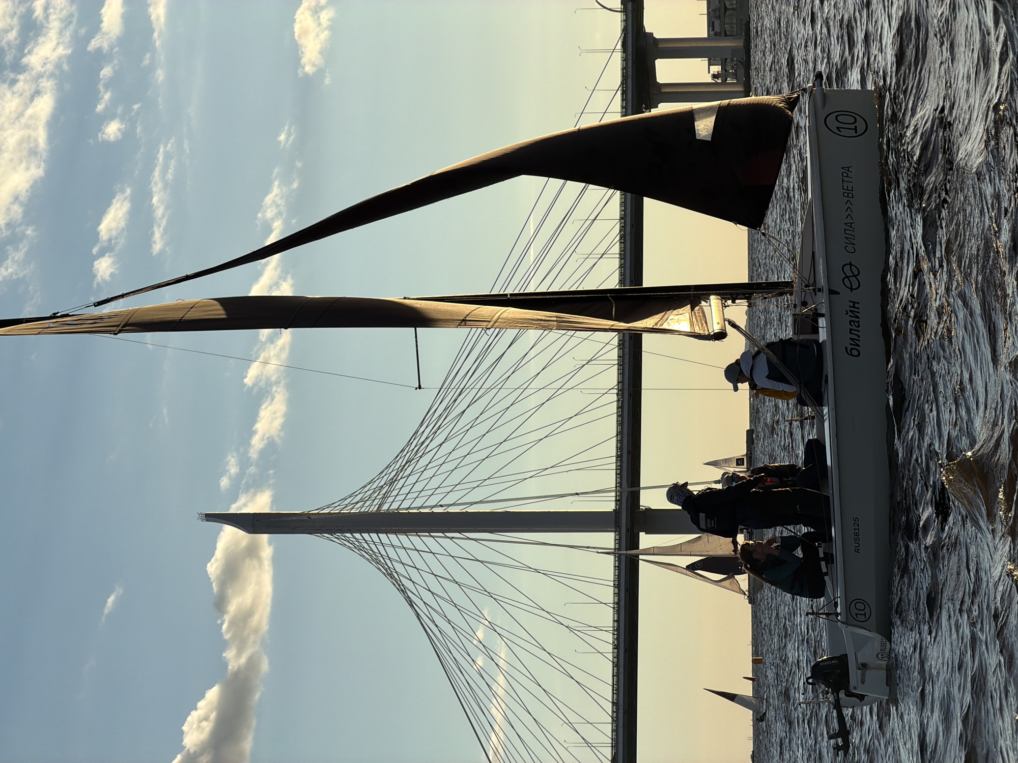 The image depicts a fleet of sailboats navigating on a body of water, with a prominent cable-stayed bridge, likely the Western High-Speed Diameter, in the background. The sky is a pale blue with scattered white clouds, and the sunlight suggests it is late afternoon or early evening. The main subject is a white sailboat in the foreground, carrying at least four individuals. One person stands at the stern, wearing dark clothing and a cap. Another person, wearing a cap and a vest, is seated near the front. Two other individuals, one seated and one crouching, are visible on the port side. The sailboat has a dark sail and bears the inscription "Билайн СИЛА>>>ВЕТРА" and the number "10" on its hull. Other sailboats, with their sails partially visible, are scattered across the water in the middle ground and background. The water is choppy, indicating a moderate breeze. The overall scene conveys a sense of leisure activity and the vastness of the urban infrastructure.