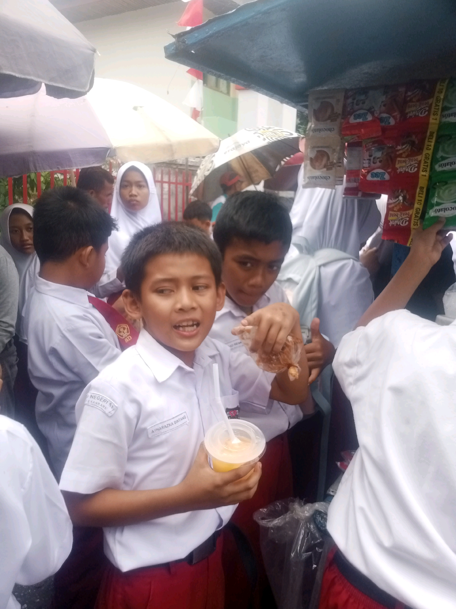 This is a photograph taken outdoors on an overcast day, likely in Pekanbaru, Indonesia, as evidenced by the "NEGERI 51 PEKANBARU" inscription on a boy's uniform. The scene depicts a group of children, appearing to be students, gathered around a street vendor's cart. The vendor's cart is laden with various packaged snacks and drinks, some of which are visible as colorful wrappers. In the foreground, a young boy in a white school uniform with red shorts is holding a clear plastic cup filled with a yellow beverage and a straw, and in his other hand, he is holding a small plastic bag containing snacks. He is looking towards the left of the frame with an open mouth, appearing to be speaking or exclaiming. Another boy, also in uniform, stands behind him to the right, holding a thumbs-up gesture and a plastic bag of snacks. Several other children, some wearing headscarves, are visible in the background, and the scene is partially obscured by umbrellas and the vendor's cart canopy. The overall atmosphere suggests a typical street scene where students are purchasing snacks from a local vendor.