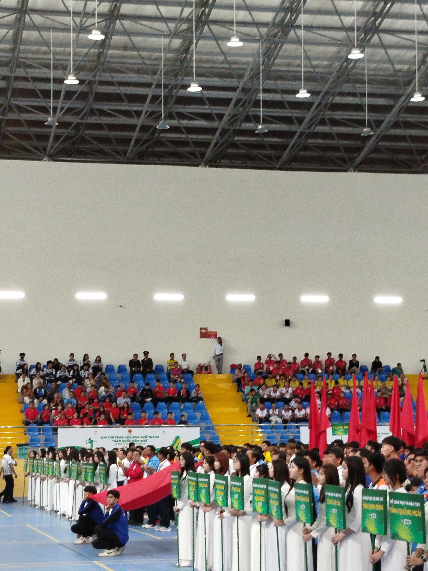 The image shows a wide shot of an indoor stadium event, likely a competition or ceremony. A large crowd is seated in tiered blue bleachers in the background, some wearing red and yellow. In the foreground, a group of young women, dressed in white áo dài, are lined up holding green signs with Vietnamese text. Red flags are prominently displayed in front of them. The stadium has a high ceiling with exposed metal trusses and hanging lights. A banner with Vietnamese text is visible behind the women. A few individuals are also visible standing on the sidelines. The image has a slightly grainy texture, suggesting it was taken with a mobile device. The presence of the banner with "GIẢI THỂ THAO HỌC SINH PHỔ THÔNG TOÀN QUỐC NĂM 2020" (National High School Sports Festival 2020) and the specific text on the green signs "SỞ GIÁO DỤC VÀ ĐÀO TẠO TỈNH QUẢNG NGÃI" (Department of Education and Training of Quang Ngai Province) indicates that the event is a national sports competition for high school students, with representation from Quang Ngai province. Da Lat is a city in Vietnam known for hosting national sports events, making it a plausible location for such a competition.