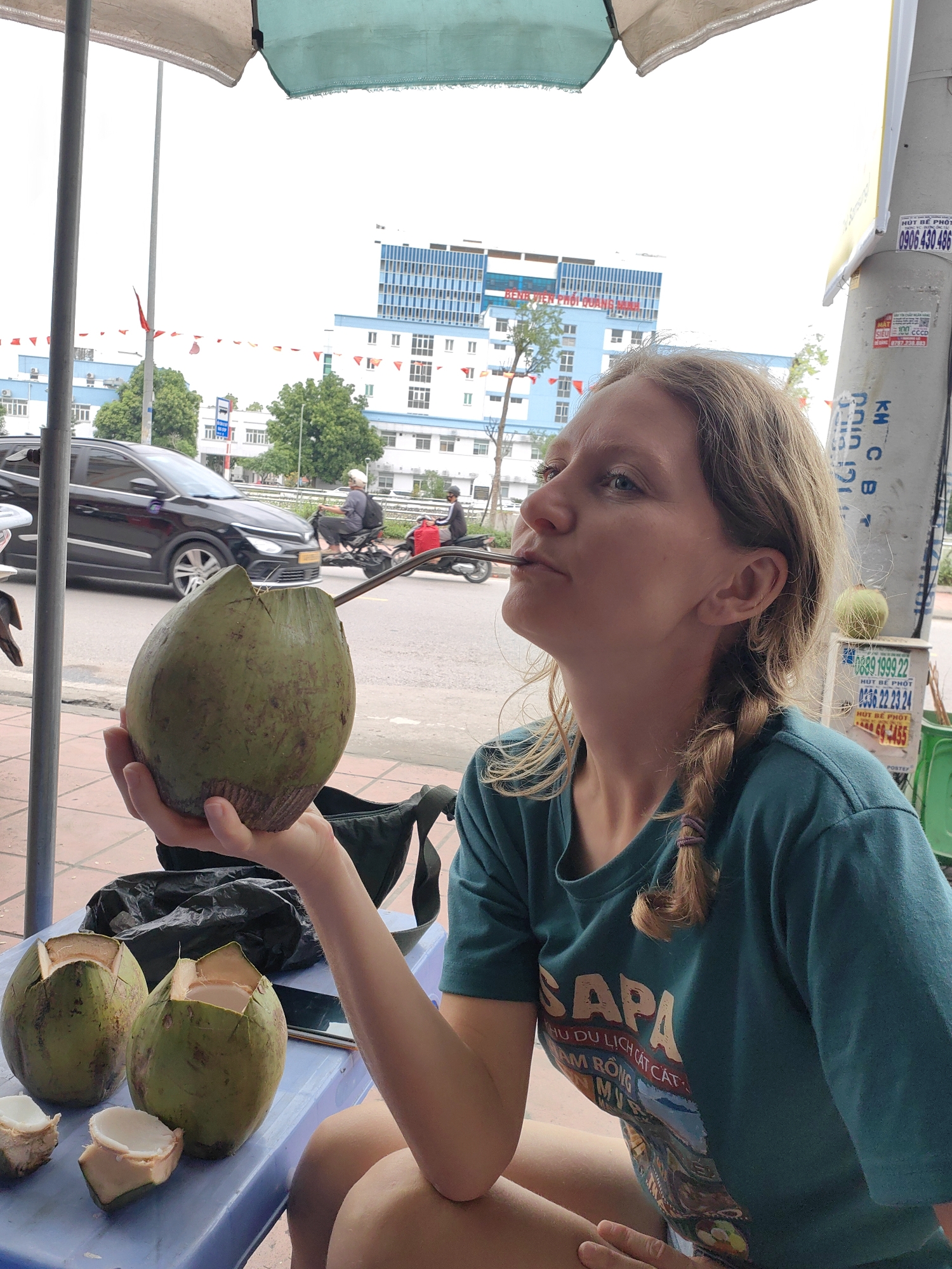 A Caucasian woman with blonde hair in a braid is drinking from a green coconut through a straw. She is sitting at a blue plastic table with three other coconuts that have been cut open. In the background is a street scene with a black car, motorbikes, and a hospital or large building flying Vietnamese flags. A sign on a pole has Vietnamese writing. The writing on the woman's t-shirt says "Sapa." The image appears to be taken in the Cao Xanh Ward of Vietnam.