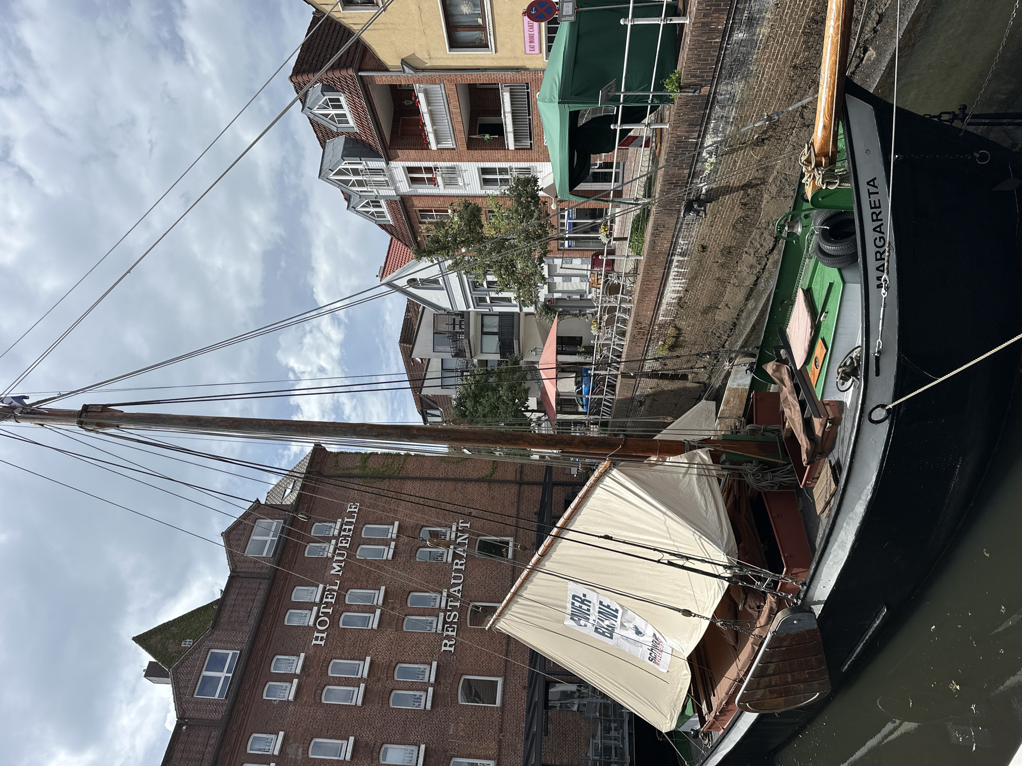 The photograph depicts a historic sailing ship named "Margareta" docked in a canal within the city of Buxtehude, Germany. The vessel's black hull is visible, with a green deck and a wooden mast extending upwards. One sail is unfurled, featuring an advertisement. The ship is docked beside a brick building with numerous windows and the sign "Hotel Muehle Restaurant" vertically printed on the façade. Other multi-story buildings line the canal on the opposite bank, some with balconies. The sky is overcast with visible cloud cover. Utility wires extend across the scene from the buildings. The boat is docked right on the edge of the canal.