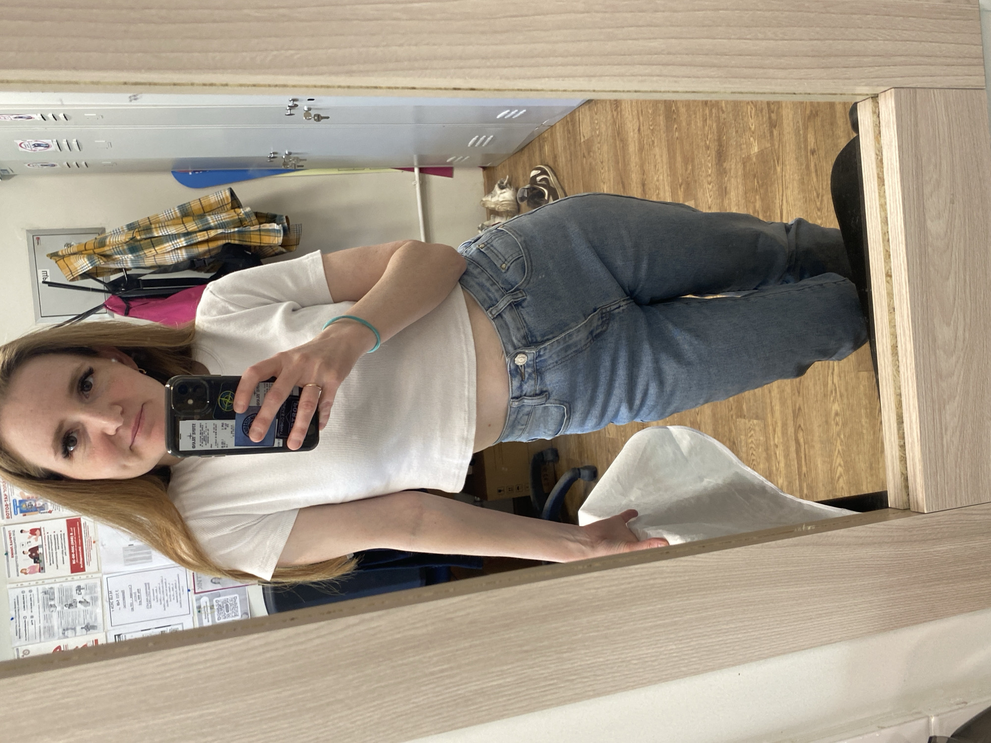 A woman is reflected in a mirror while taking a selfie in what appears to be a locker room or storage area. The location might be Chaikovsky, Russia. The woman has light skin and long light brown hair. She is wearing a white cropped t-shirt, jeans, and a thin blue bracelet. In her right hand, she holds a smartphone with a dark case, decorated with stickers on the back. The background shows light gray lockers with some keys in the locks, a wooden floor, and a white wall covered in documents. A yellow plaid garment is draped on top of some of the lockers. In the right foreground, the frame of the mirror is visible.