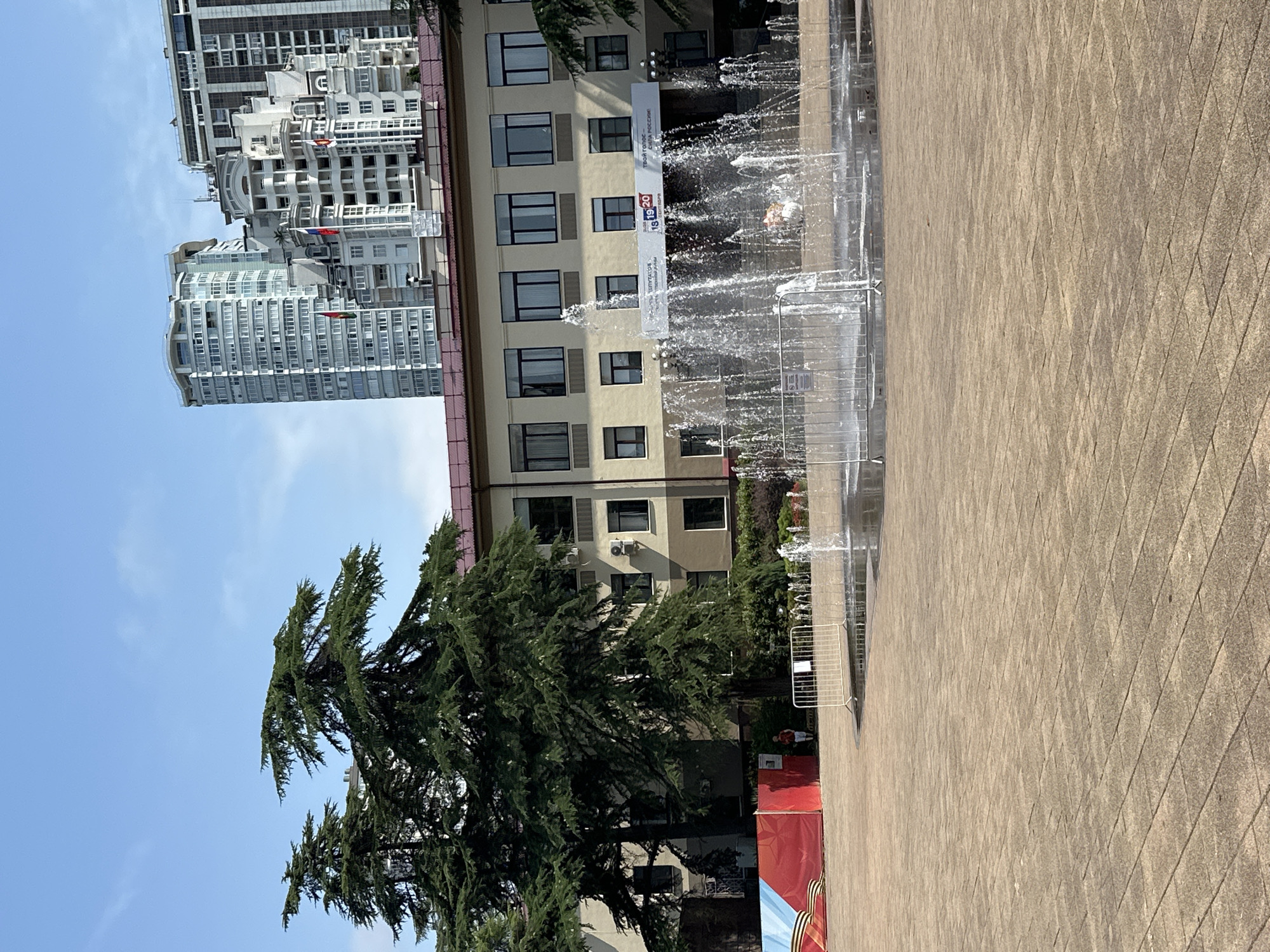 A public square in Sochi, Russia, features a multi-tiered fountain in the foreground with water jets spraying upwards. The square is paved with large, light-colored square tiles. In the mid-ground, a low white metal fence surrounds part of the fountain area. Behind the fence, a light yellow building with dark-framed windows is visible, with a banner displaying the numbers "18 19 20" and text in Russian. Large evergreen trees with dark green foliage are situated to the left of the building, partially obscuring it. To the far left, a red banner with blue and white elements is displayed on a temporary structure. In the background, several tall, modern apartment buildings with numerous windows dominate the skyline under a clear blue sky. A few people are visible in the distance, near the red banner.