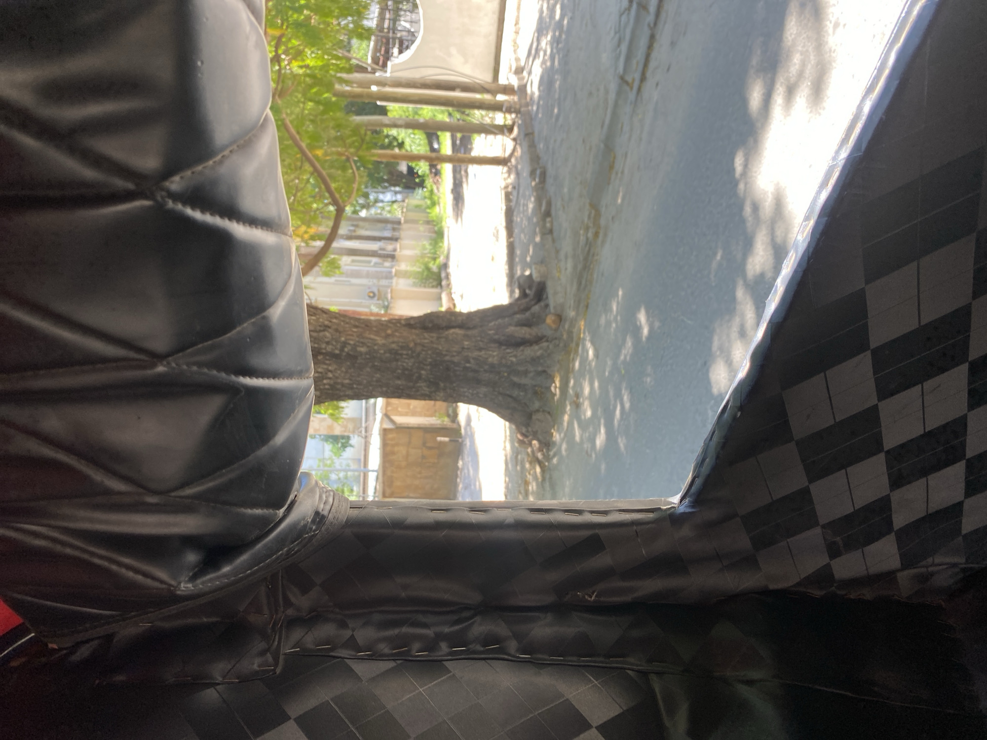 The image is taken from inside a vehicle, possibly a rickshaw or auto-rickshaw, looking out onto a street scene. The foreground is dominated by the interior of the vehicle, showing parts of the padded seating and the frame's covering material, which has a checkered pattern.
Outside, the view includes a paved street, trees, and buildings. Sunlight filters through the leaves, creating dappled shadows on the road. A large tree trunk is visible on the side of the road.
Without additional information, pinpointing the exact location in Dar es Salaam, Tanzania, is not possible.