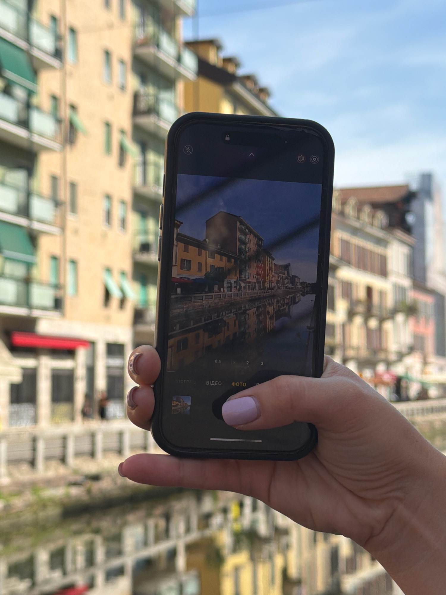 A person's hand, with light skin and a pastel purple fingernail polish, holds an iPhone in portrait orientation. The phone's screen displays a reflection of buildings along a canal, captured in what appears to be Milan, Italy, likely along the Naviglio Grande or Naviglio Pavese due to the distinctive architecture and waterway. The background shows a blurred view of similar buildings with balconies and awnings, as well as a bright blue sky. The phone is positioned in the foreground, partially obscuring the view of the canal and buildings behind it. The focus is on the hand and phone, with the background and the image on the phone's screen providing context.