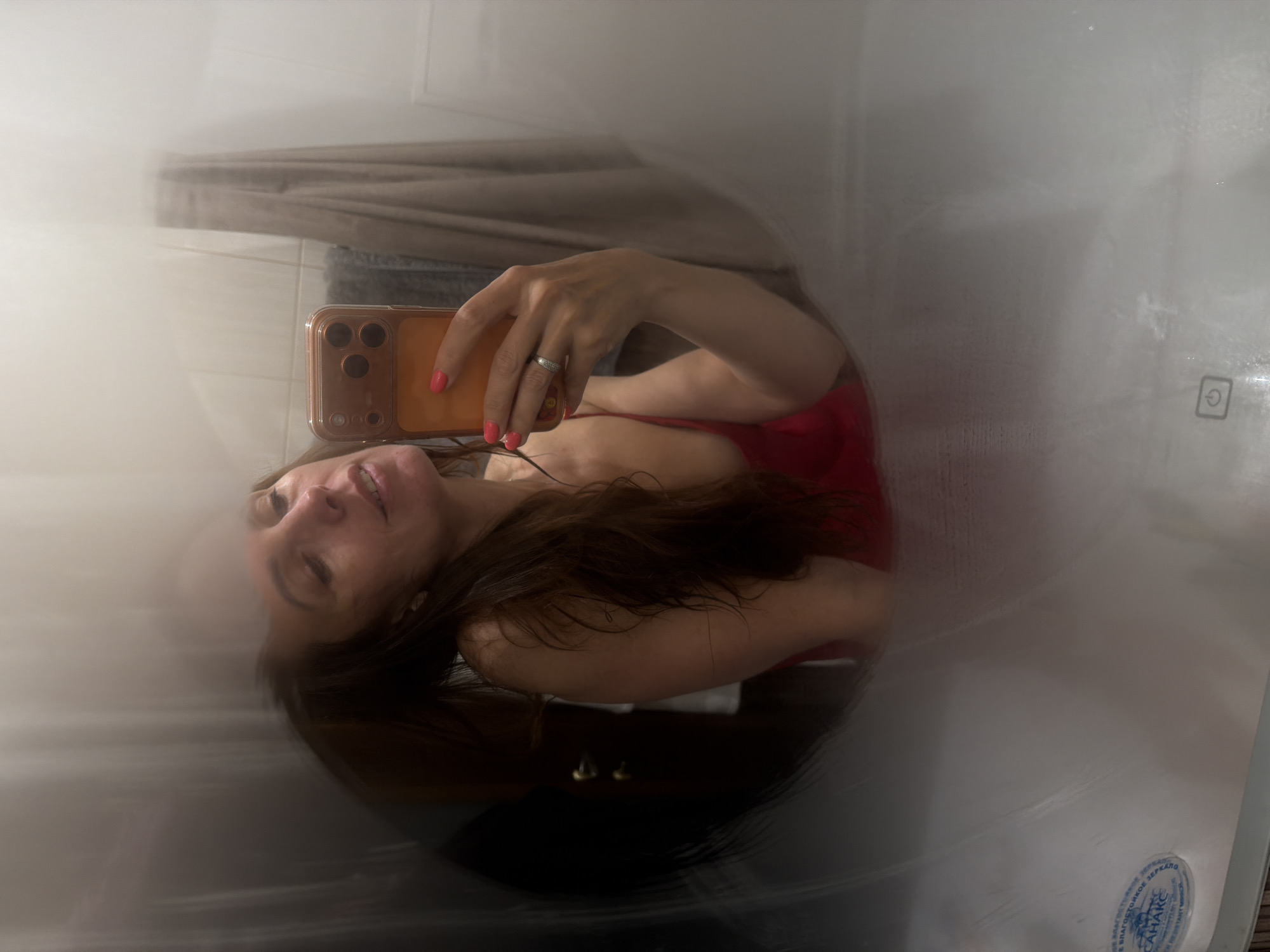 The image is a tilted selfie of a woman, taken through a translucent or frosted surface, possibly a shower screen or a steamy mirror, which creates a soft, diffused effect with visible streaks.
**Subjects:** The central subject is a woman with long, dark brown hair, captured from the chest up. She wears a red top or swimsuit and has her nails painted a bright coral or reddish-orange. A textured silver ring is visible on her left ring finger. She holds a light brown or amber-colored smartphone, equipped with a multi-lens camera array, and has white earphones plugged into it, running towards her ear.
**Scene and Setting:** The background suggests a bathroom. Faintly visible behind her is a beige or brown shower curtain and a darker-colored towel. To the right, a smooth, white surface, likely a bathtub or shower wall, occupies a significant portion of the frame.
**Activity:** The woman is taking a selfie, holding her smartphone to capture her reflection or image through the translucent barrier. She has a slight smile and is looking towards the phone.
**Notable Details:** The image's soft focus and tilted perspective give it a candid and somewhat intimate atmosphere. The lighting appears to be indoor artificial light. The diffusion from the foreground element creates a dreamlike quality.
**Visible Text:** On the white surface to the right, a universal power symbol (a circle with a vertical line) is clearly visible. Below it, a circular blue and white label contains Cyrillic text, including "ЗАЩИЩЕННОЕ ЗЕРКАЛО" (Protected Mirror), "ВЛАГОСТОЙКОЕ" (Moisture-resistant), and the brand name "САНАКС" (SANAX). The English words "RESISTANT MIRROR" are also part of this label, indicating a product made for Russian-speaking markets, consistent with the given location context of Birobidzhan, Russia.