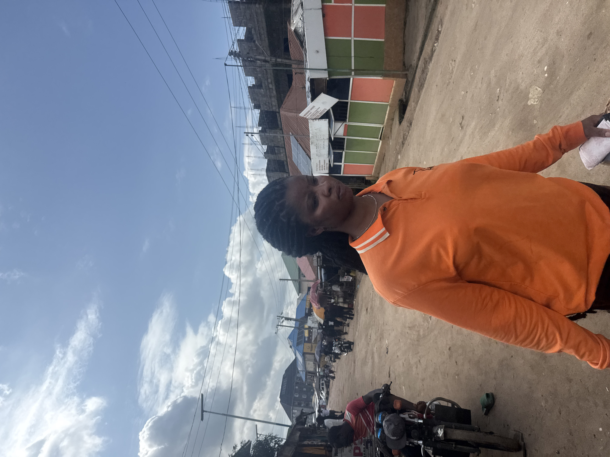 A person in an orange long-sleeved shirt stands in a street scene in Ihiagwa, Nigeria. The sky is blue with scattered white clouds, suggesting it is daytime. The ground is a dirt and gravel road, and buildings are visible in the background. Some buildings are under construction, with scaffolding and exposed concrete. Others have colorful panels on their facades. Power lines crisscross the scene.
In the foreground, a motorcycle is parked on the right. The person in the orange shirt is looking towards the camera with a neutral expression. Their hair is styled in braids. Another person wearing a red and white striped shirt is partially visible, leaning over the motorcycle.
Further down the street, there are more buildings, some with awnings or tarps, and several people and motorcycles. The overall impression is of a busy, active street in a developing area. The time of day appears to be midday, given the bright sunlight and clear sky. There is no discernible text in the image.