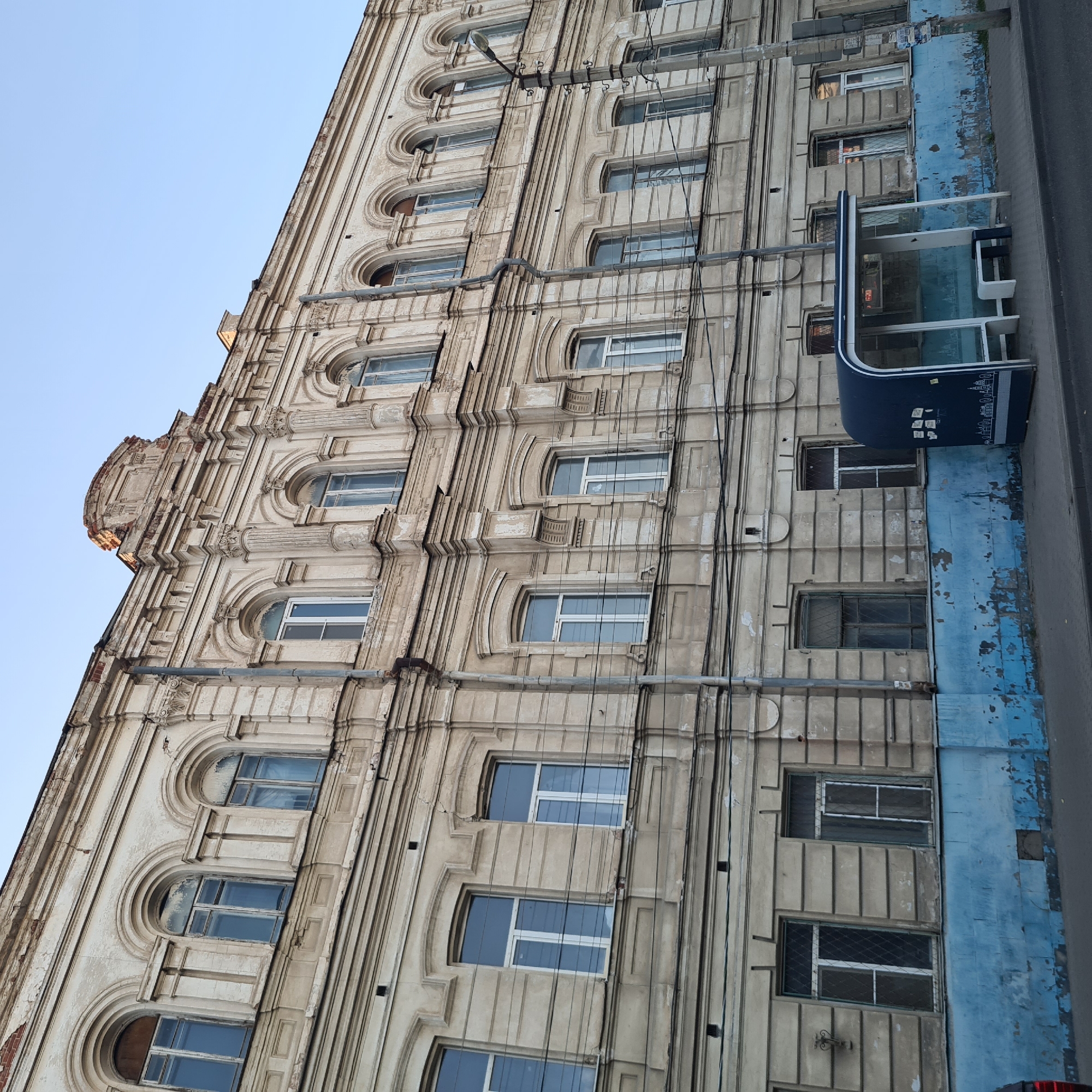The image depicts a street scene in Astrakhan, Russia, featuring an old, cream-colored building with an ornate facade and multiple arched windows. The building shows signs of age, with some peeling paint and cracks. Below the main building structure, there is a bright blue band of paint, also with visible wear.
In the foreground, to the right of the frame, is a modern, dark blue bus stop shelter with glass panels. Inside the shelter, a white bench is visible, along with a dark trash receptacle. The bus stop shelter is decorated with a white silhouette of city buildings.
A concrete lamppost with multiple wires attached stands to the right of the bus stop. The street in front of the bus stop appears paved, and a portion of a vehicle, possibly red, is visible on the far left edge of the image. The sky above is a clear, pale blue, suggesting it is daytime. The overall scene conveys a sense of urban life with a juxtaposition of old architecture and modern infrastructure.