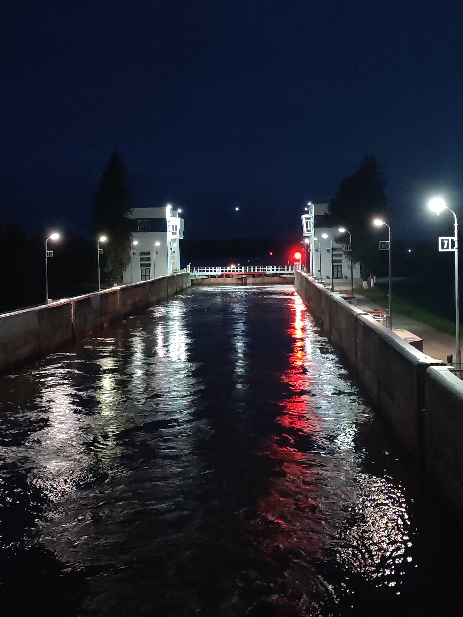 The image depicts a lock on a body of water at night in Belousovo, Russia. The lock walls are made of concrete and are illuminated by streetlights and red navigation lights. The water surface is reflecting the lights, creating a shimmering effect. In the background, there are buildings with windows and more lights. The sky is dark blue and clear. There are no people visible in the image.