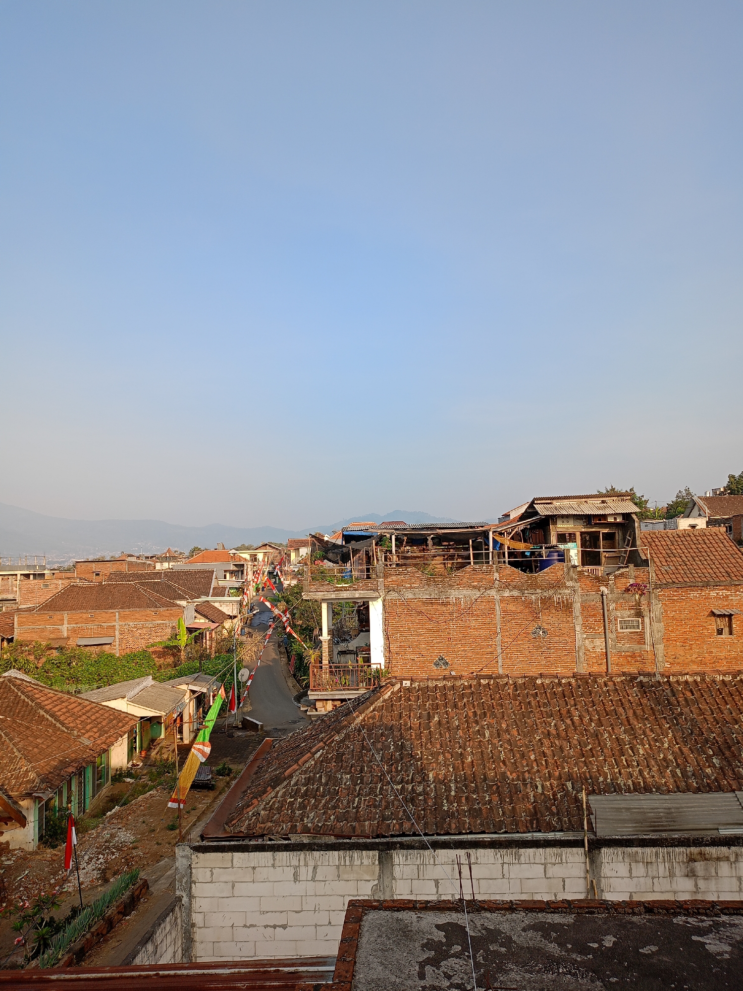 The image captures a view of a residential area in Batu, Indonesia, likely from an elevated position. The scene is dominated by houses with red tile roofs, constructed primarily from brick. A paved road winds through the neighborhood, lined with more houses and vegetation. Several Indonesian flags, red and white, are visible, suggesting a national holiday or celebration. The sky is clear and blue, with distant hills or mountains faintly visible on the horizon. The overall impression is that of a quiet, possibly rural or semi-urban, community during the daytime.