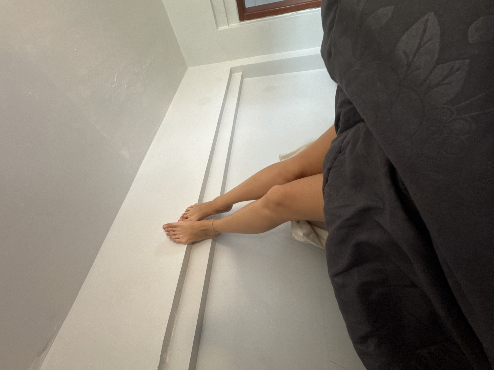 In North Kuta, Indonesia, a low-angle medium shot shows a person's bare legs and feet resting against a white wall. The person is lying down, with their legs extended upwards. The wall has two horizontal bands, one grey and one white. Below the wall, dark fabric, possibly a blanket or duvet, covers the person's lower body. The lighting suggests it is daytime, and the overall environment appears to be a bedroom.
