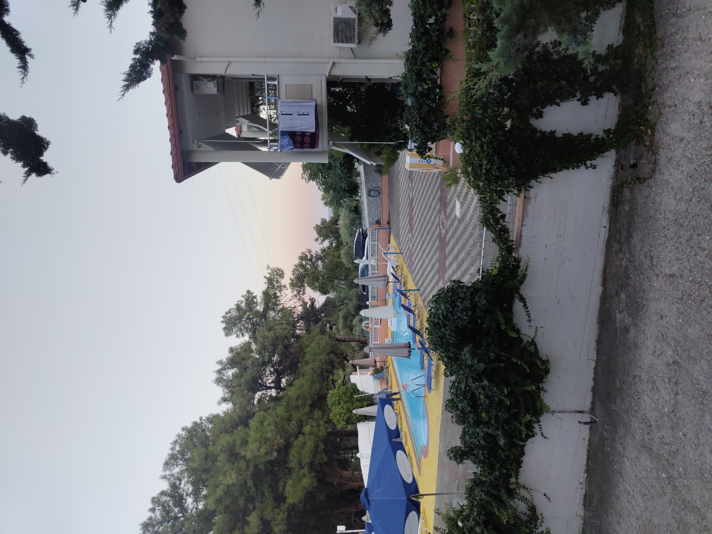 The image captures a resort-like setting, likely in Limenaria, Greece, featuring a swimming pool area. A light blue swimming pool with a yellow deck is visible, surrounded by lounge chairs and umbrellas. To the right, a multi-story building with a balcony displays several towels, suggesting it's accommodation. Lush green trees frame the scene, and a white wall covered in ivy forms the foreground, hinting at a private or hotel property. The sky is overcast with a soft gradient, indicating early morning or late evening. The presence of lounge chairs and a pool implies a leisure or hospitality venue.