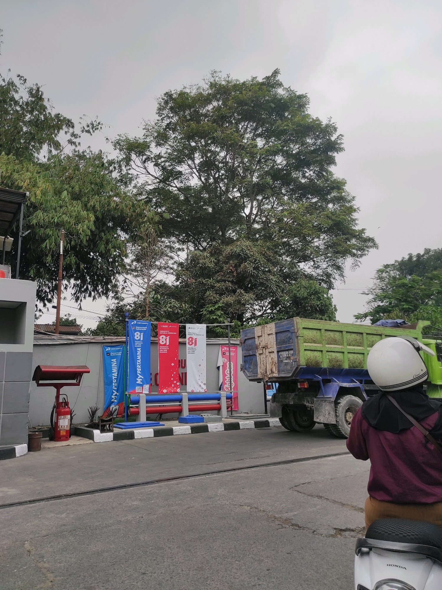 In Padalarang, Indonesia, a view captured from a scooter shows a Pertamina gas station. In the foreground, a person wearing a maroon jacket and tan pants rides a white Honda scooter. Behind them, a large green dump truck is parked. Several vertical banners, featuring the "81" logo and the "MYPERTAMINA" text, stand in front of a grey wall. A red fire extinguisher is visible next to a gas pump. Lush green trees and an overcast sky form the backdrop.
