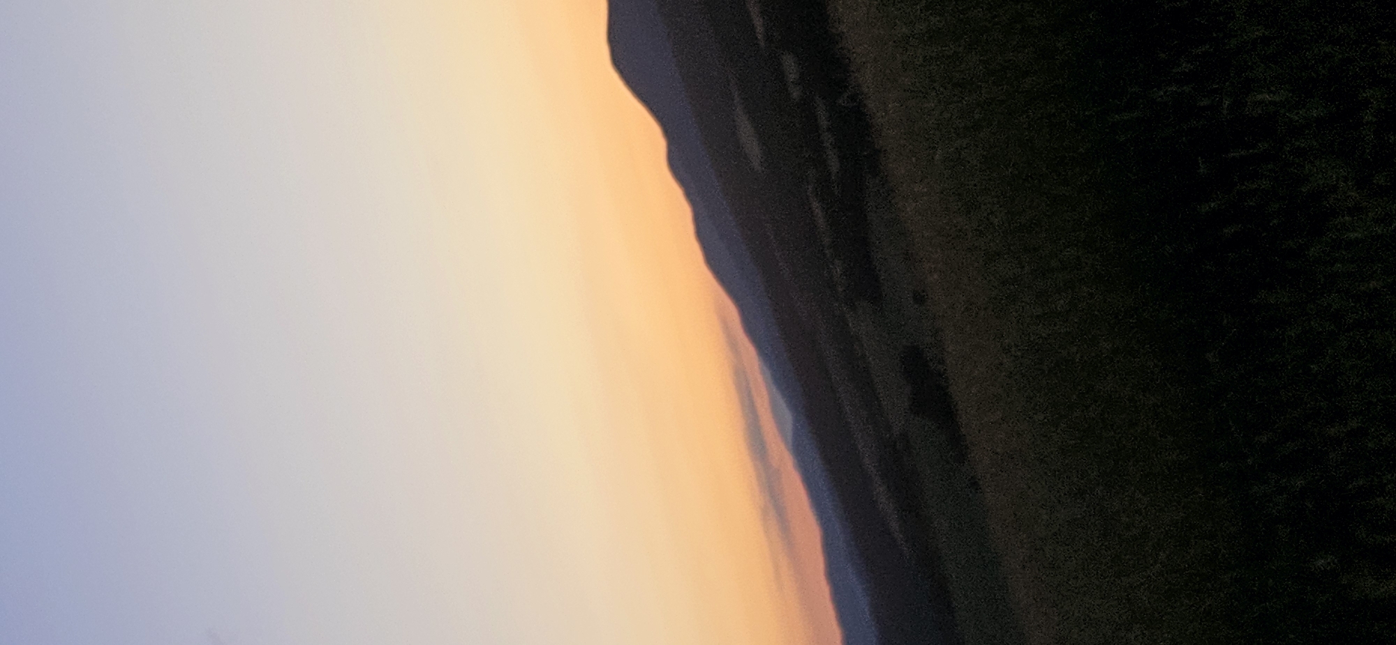 The image depicts a landscape scene at dusk or dawn. The sky transitions from a pale lavender at the top to a warm orange hue towards the horizon. Silhouetted against the colorful sky are rolling hills or mountains, with darker layers of hills receding into the distance. The foreground is dominated by a dark, grassy field. The overall impression is one of a natural, open environment.