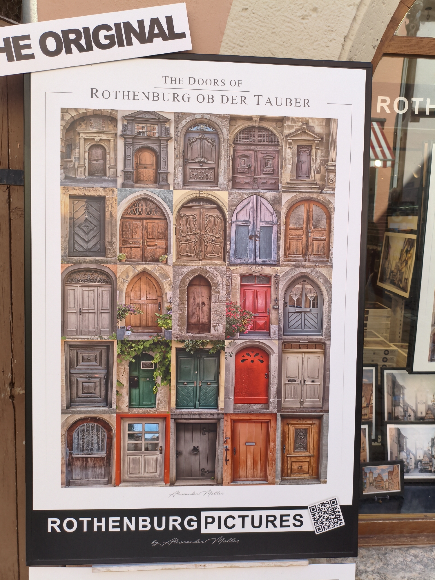 The image is a photograph of a poster displayed on a wooden surface. The poster features a collage of 24 photographs of doors, arranged in a 4x6 grid. The title of the poster reads "THE DOORS OF ROTHENBURG OB DER TAUBER," indicating the location as Rothenburg ob der Tauber, Germany. The photographs showcase a variety of architectural styles and colors of doors, with some featuring intricate carvings and ornate details. The bottom of the poster has the text "ROTHENBURG PICTURES" with the artist's name "by Alexander M ller" printed below. A QR code is also visible in the bottom right corner of the poster. In the background, a shop window is visible, displaying more framed pictures and architectural elements. The overall scene appears to be an outdoor display, possibly in a tourist area of the city.