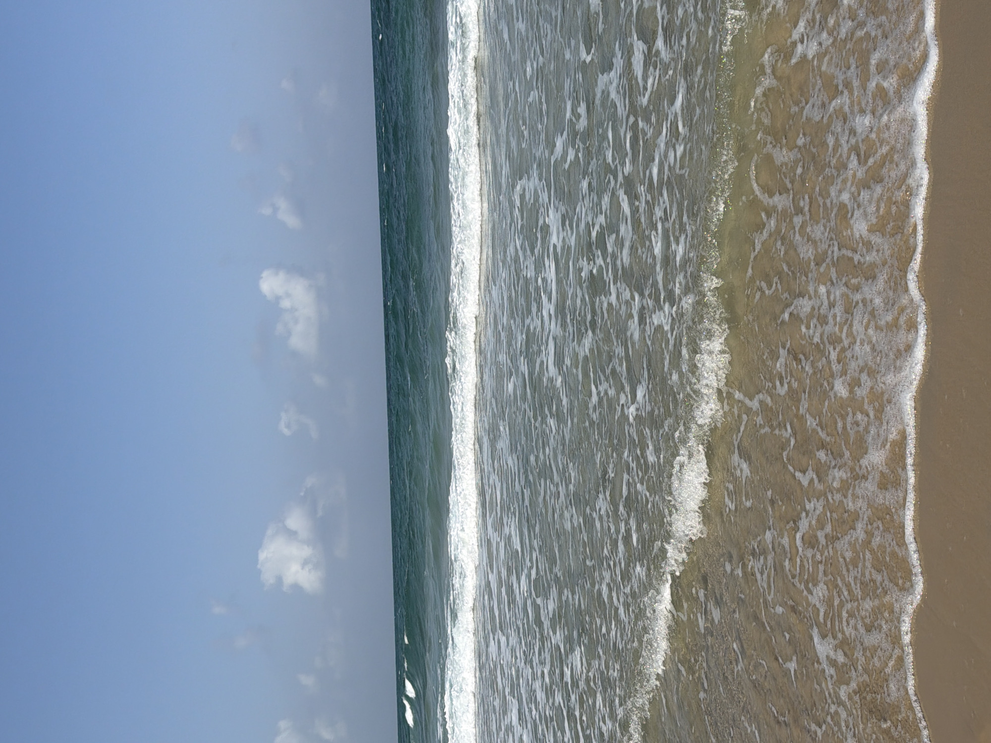 The image depicts a view of the Mediterranean Sea from a beach in Bat Yam, Israel. The sky is clear blue with a few scattered white clouds. The ocean is a deep turquoise color with white waves breaking near the shore. The foreground is a sandy beach with the edge of a gentle wave lapping at the sand. There are no people or man-made objects visible in the frame. The scene is one of natural coastal beauty.