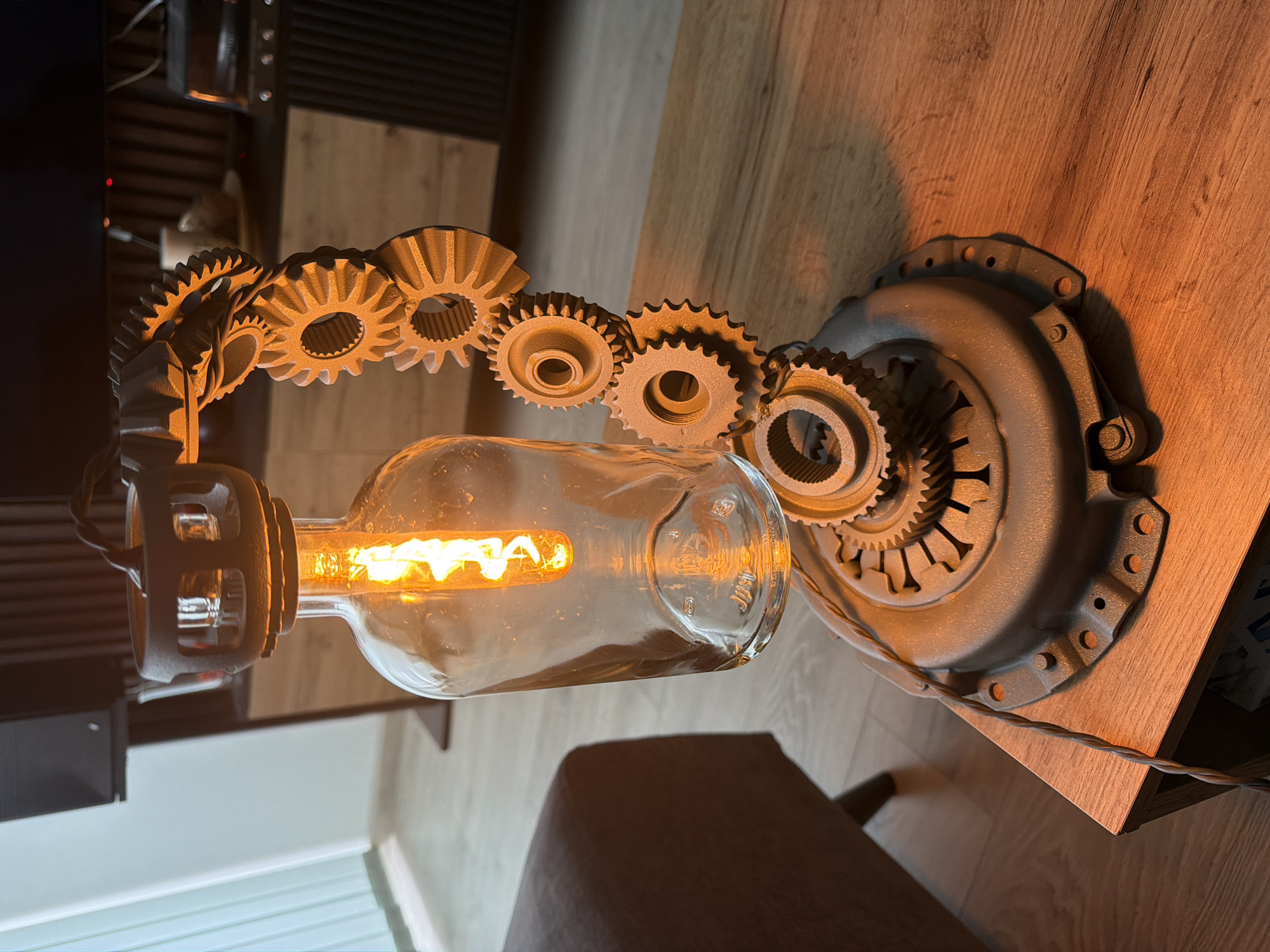 An illuminated, industrial-style lamp made from repurposed mechanical components is prominently displayed on a wooden surface. The lamp's base appears to be a grey, circular car clutch assembly, from which an array of gears extends upwards and outwards. These gears, varying in size and design, are arranged in an arc, creating a decorative and functional structure. A clear glass light bulb with a visible, glowing Edison-style filament is suspended within this arrangement, emitting a warm, orange light that casts long shadows and highlights the textures of the metal and wood.
The wooden surface, likely a table or shelf, has a light to medium brown tone with visible grain. In the blurred background, hints of other indoor furniture, possibly a dark chair or sofa, and a dark wall unit or speaker system are visible. The overall lighting suggests an indoor setting, possibly during the evening, where the lamp provides the primary illumination.
On the glass light bulb, near its base, some embossed text is visible, including what appears to be "JUST" along with other symbols and numbers, possibly a CE mark and manufacturing codes like "21724." The lamp's design showcases a strong steampunk or upcycled aesthetic.