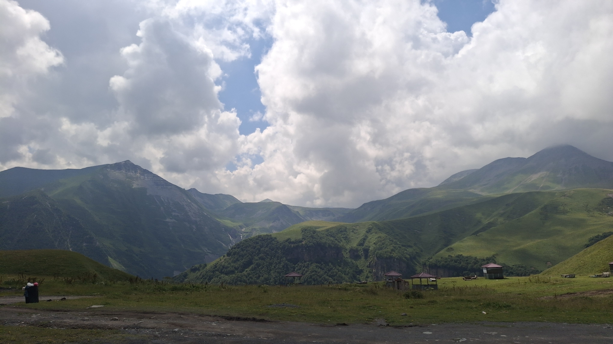 The image shows a mountainous landscape with lush green slopes under a cloudy sky. In the foreground, a dirt road leads to a grassy area with several picnic shelters. To the left, a person stands beside a black trash bin, with red and white bags inside. The background features several peaks, some with rocky faces and others covered in vegetation. A small waterfall can be seen cascading down a mountainside in the distance. The scene is serene and depicts a natural environment, possibly a tourist or recreational area in Gudauri, Georgia, known for its mountainous terrain and outdoor activities.