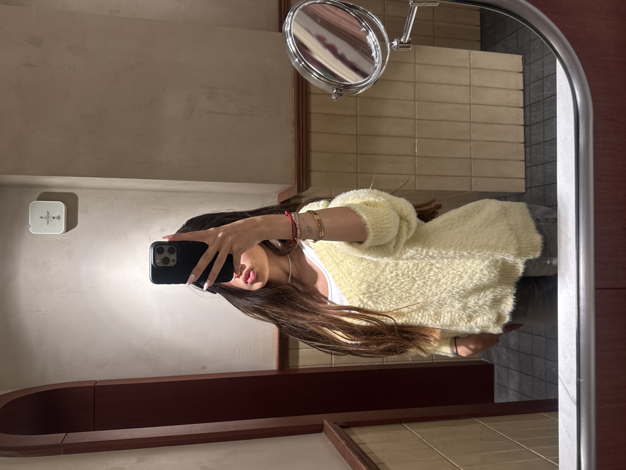The image displays a mirror selfie taken by a person in what appears to be a modern restroom or vanity area located in Yerevan, Armenia. The individual, seen from the chest up, holds a black-cased smartphone with their left hand, partially obscuring their face, with only their lips, chin, and lower nose visible. They are wearing a soft, light yellow fuzzy cardigan over a white top, and their long, dark brown hair cascades down their shoulders. Their visible right arm features an assortment of bracelets, including a red string bracelet and several gold-toned bangles, one notably adorned with an "evil eye" charm.
The background reflects a contemporary design with light beige rectangular subway tiles on the upper wall and darker grey tiles on the lower sections. A circular, chrome-framed magnifying mirror on an articulated arm is mounted on the wall above and to the right of the person's head. To the left, a small, white square device, possibly a sensor or control panel, is affixed to the wall, displaying a faint icon (resembling a leaf or flame) and some minute, illegible text. A dark brown, possibly wooden, panel or shelf runs horizontally across the mid-section of the reflection. The indoor lighting suggests no specific time of day.