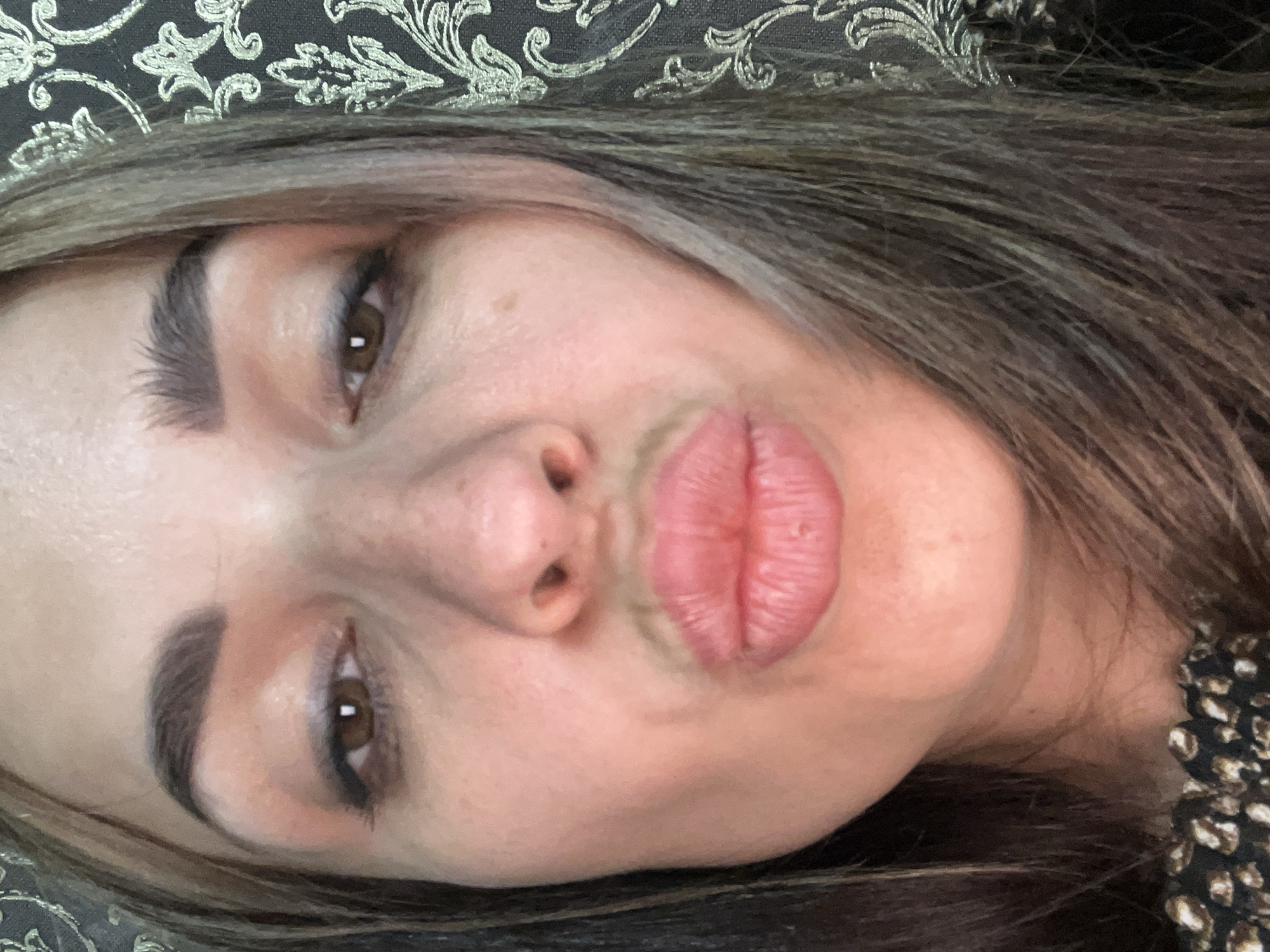 A close-up selfie shows the face of a woman with brown hair and brown eyes. She has her lips pursed in a kissing gesture. Her eyebrows are dark and well-defined. She is wearing subtle makeup, with some eyeshadow visible on her eyelids. The background is a dark, patterned wallpaper with a floral design in a lighter color.
