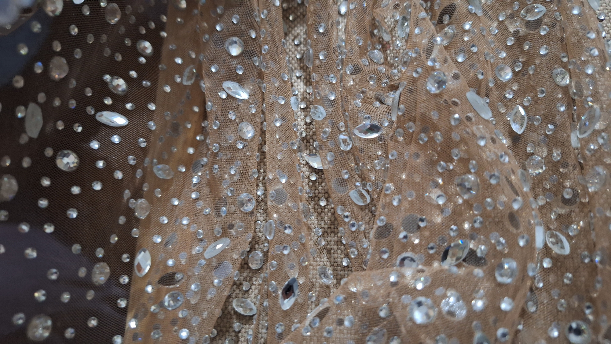 The image shows a close-up of a light brown, semi-transparent fabric covered with numerous clear, reflective crystals or rhinestones of varying sizes and shapes. The fabric appears to be draped or gathered, creating folds and shadows that add depth to the image. Some sections reveal a textured, possibly beaded, layer beneath the sheer fabric. The density of the crystals varies across the fabric.
There is no indication of the location being Blagoevgrad, Bulgaria or any specific urban environment; the image focuses solely on the fabric and its embellishments. No people, actions, or interactions are visible in the image.