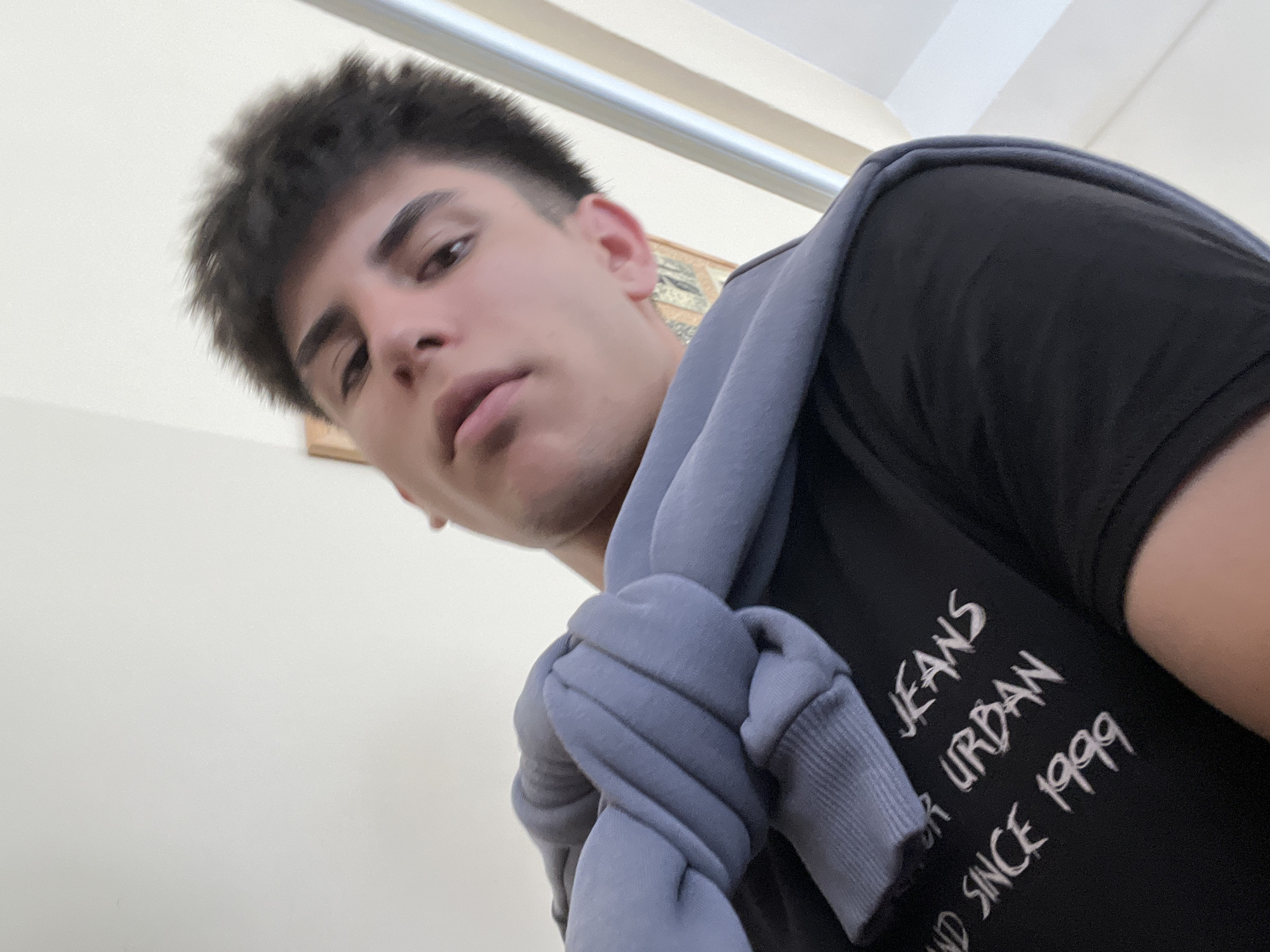 This is a close-up, low-angle selfie of a young man with dark, styled hair. He is wearing a black t-shirt with white text that reads "JEANS URBAN AND SINCE 1999" and has a light blue sweatshirt draped over his shoulders. The background is a plain, light-colored wall with a vertical bar on the right side and a framed picture visible on the left. The image is taken indoors, likely in a residential setting, and does not contain any identifiable landmarks or context that would place it in Stara Zagora, Bulgaria.