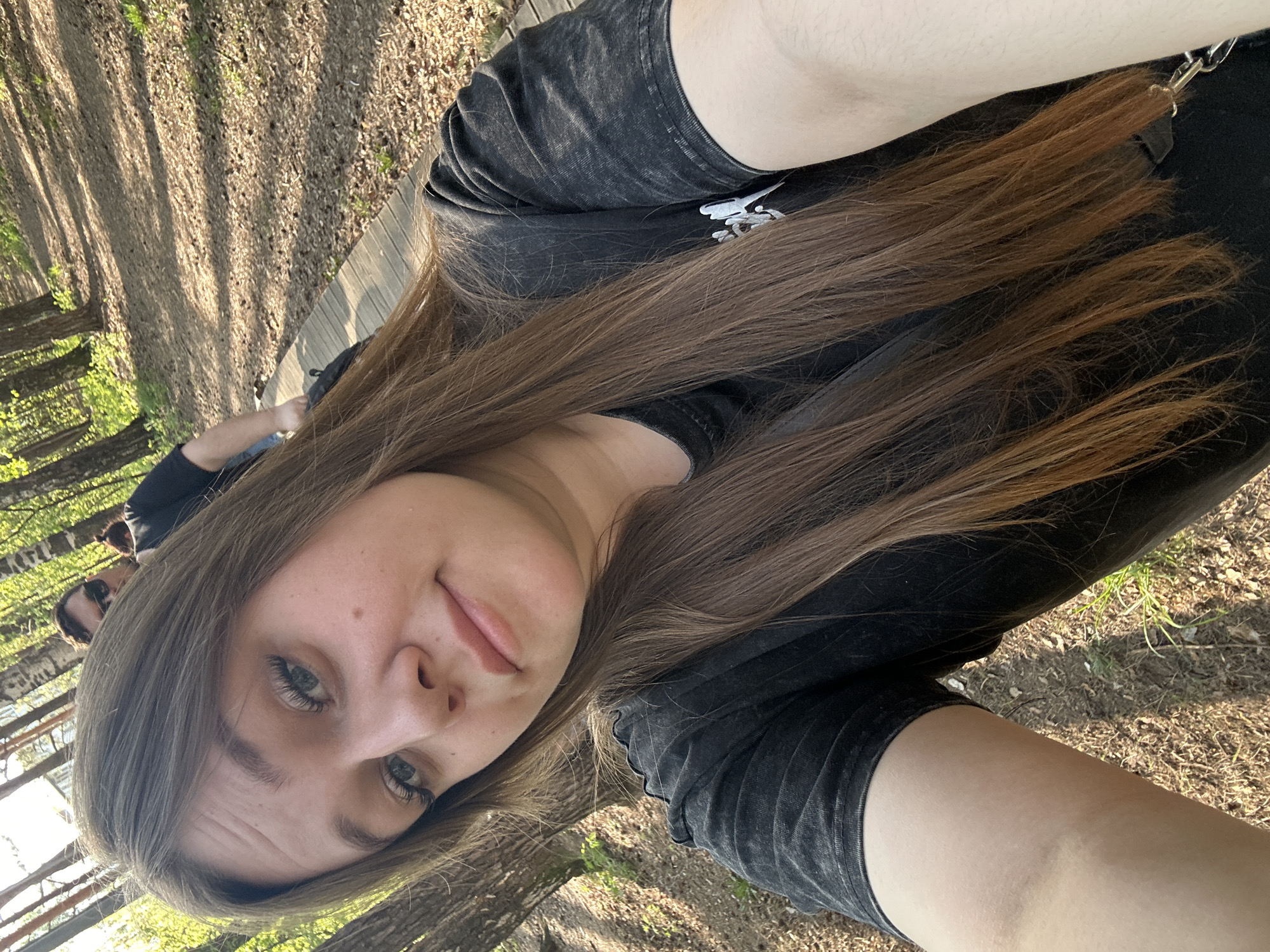 A selfie shows a young woman with long brown hair wearing a black t-shirt. The background appears to be an outdoor setting, likely a park or wooded area, evidenced by trees and ground cover. Other people can be seen in the background, although they are out of focus. The location is Chaikovsky, Russia.