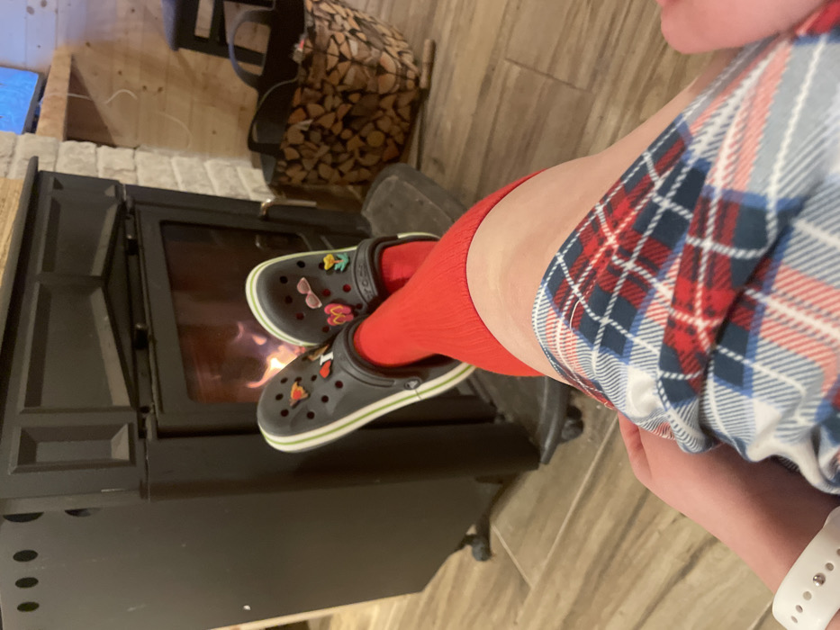 This image, reportedly located in Palež, Montenegro, displays a first-person perspective view of a person's lower body within an indoor setting. The individual's legs are visible from above the knee, clad in a red, blue, and white plaid garment, likely pajamas or lounge pants. Bright red mid-calf socks are worn on both feet, which are inserted into dark gray Crocs featuring light green and white striped soles. These Crocs are accessorized with multiple colorful Jibbitz charms, including a red heart, pink butterfly, and yellow flower, among others. A white wristband, possibly a smartwatch, is visible on the left wrist. The feet are positioned elevated, directly in front of a black, metal wood-burning stove that exhibits active flames behind its glass door. The stove rests on a dark metal floor plate. To the right of the stove, a woven basket filled with split firewood is present. The floor consists of light brown, wood-patterned tiles. The background features a wall segment constructed from light-colored stacked stone, adjacent to a section of light wood paneling. A narrow wooden shelf is visible above the stone wall, and a portion of a lighted screen, possibly a television, is discernible in the upper right corner.