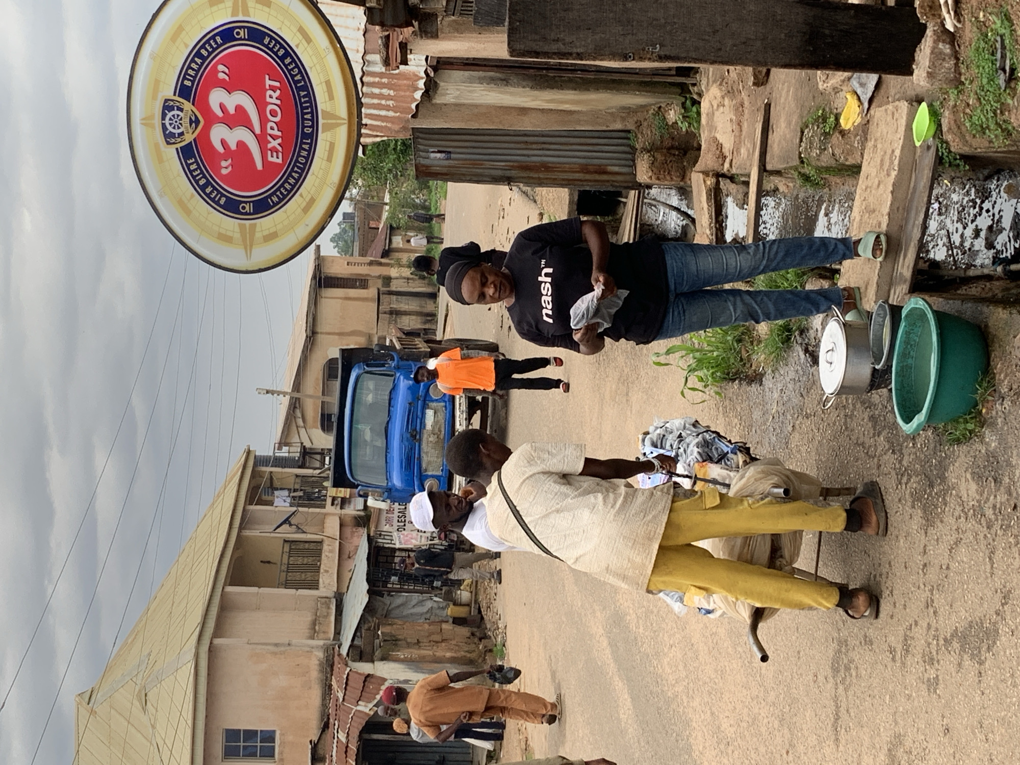 This image depicts a street scene in Jos, Nigeria. In the foreground, a person wearing yellow pants and a light-colored shirt is pushing a wheelbarrow filled with goods. To the right, a woman in a black t-shirt with the word "nash" printed on it and blue jeans is holding a piece of white cloth. Several other people are visible in the background, including a man in an orange shirt and a man in a white cap. A large blue truck is parked on the street, and a prominent circular sign for "33 EXPORT BIER BIERE" hangs overhead. The environment appears to be a commercial or residential area with buildings made of various materials, including corrugated metal. A drainage channel runs alongside the street. The sky is overcast.